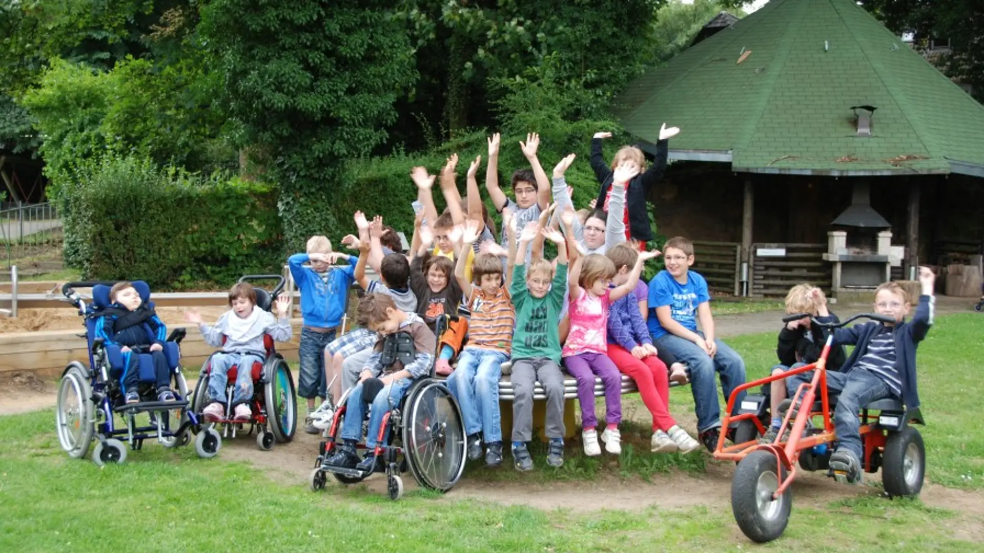 Eine Gruppe mit etwa 20 Kindern im Grundschulalter posiert für ein gemeinsames Foto an einem Spielplatz draußen im Freien. Die Mehrheit der Kinder sitzt dicht nebeneinander auf einem runden Klettertisch aus Metall. Drei Kinder mit komplexer Beeinträchtigung sind im Rollstuhl und stehen neben dem Tisch links. Zwei Schüler in Tretbuggies sind auf der anderen Seite des Tisches. Alle Kinder schauen in die Kamera und manche heben die Arme hoch in eine Art fröhliche Begrüßung.