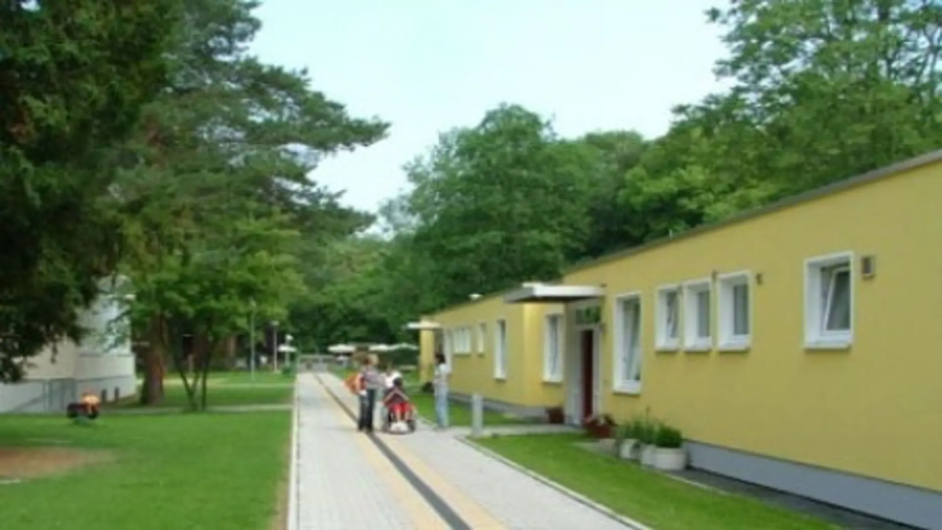 Das Internatsgebäude der LVR-Louis-Braille-Schule von draußen. Dieses befindet sich rechts im Bild. Es ist ein flacher Bau, nur mit Erdgeschoss und mit mehreren Eingängen und vielen Fenster der Zimmer. Davor ist ein breiter, betonierter Fußweg mit taktilen Markierungen zur Orientierung. Vor dem einen Eingang stehen drei Personen, die sich unterhalten, eine davon schiebt noch eine Person im Rollstuhl. Links im Bild befindet sich ein anderes Schulgebäude. Links und rechts hinter dem Internat sind viele Bäume mit grünen Baumkronen zu sehen.