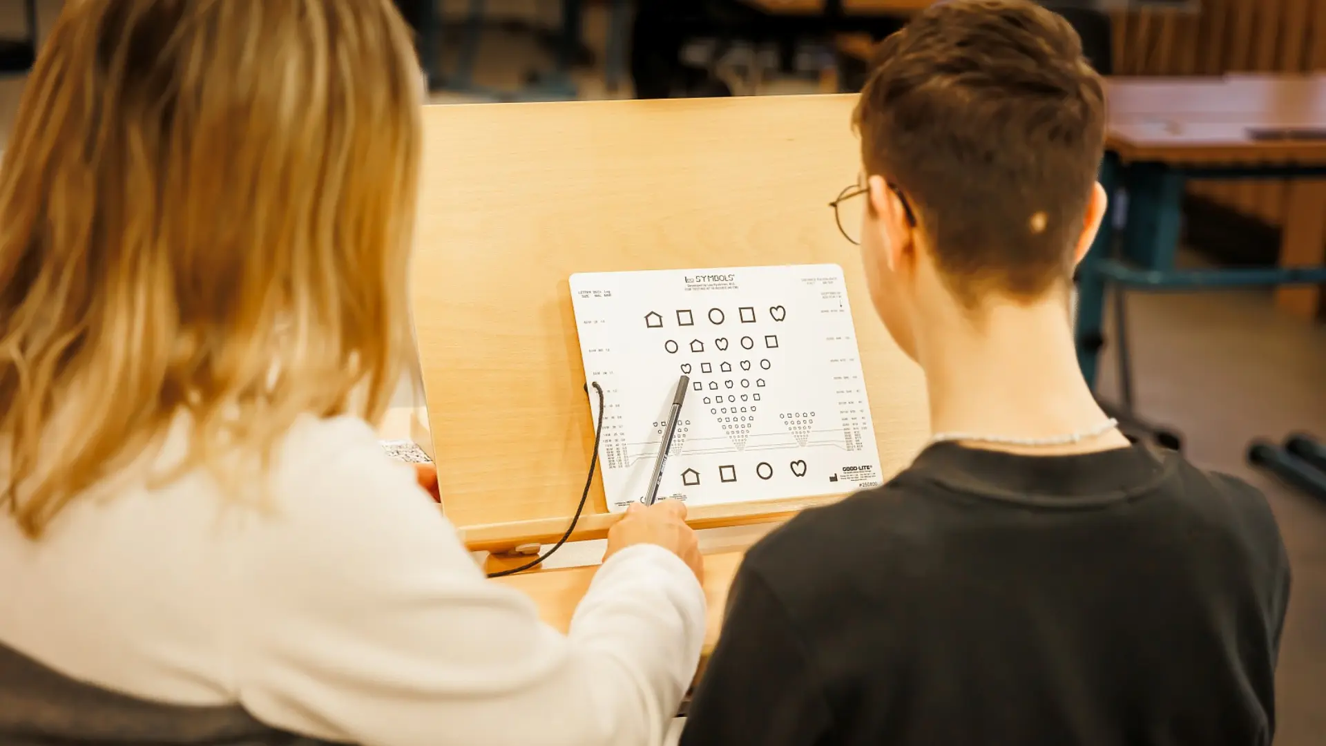  Eine Lehrkraft sitzt mit einem Schüler vor einem hochgekippten Schreibtisch. Beide blicken zu einem Blatt mit Symbolen in verschiedenen Formen und Größen, die man für die Untersuchung der Sehkraft benutzt. Das Foto wurde über ihre Schulter aufgenommen.