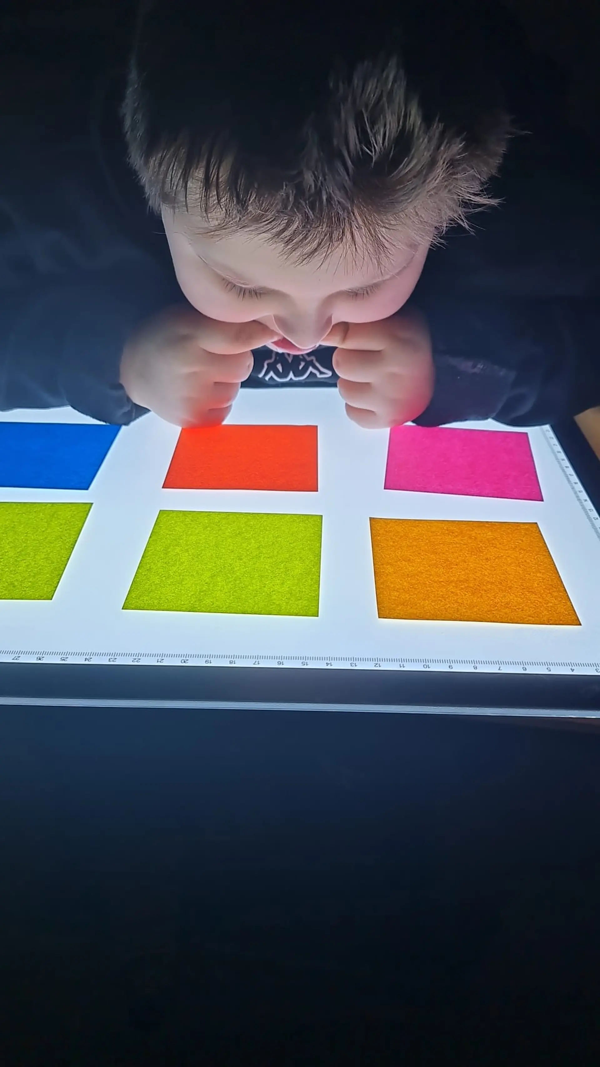 Ein Junge mit komplexer Beeinträchtigung sitzt ganz nah an einem Lichttisch. Auf der Oberfläche liegen sechs quadratische Papierblätter in verschiedenen Farben. Das Kind schaut sich diese konzentriert an und beißt dabei an seinen Zeigefingern.