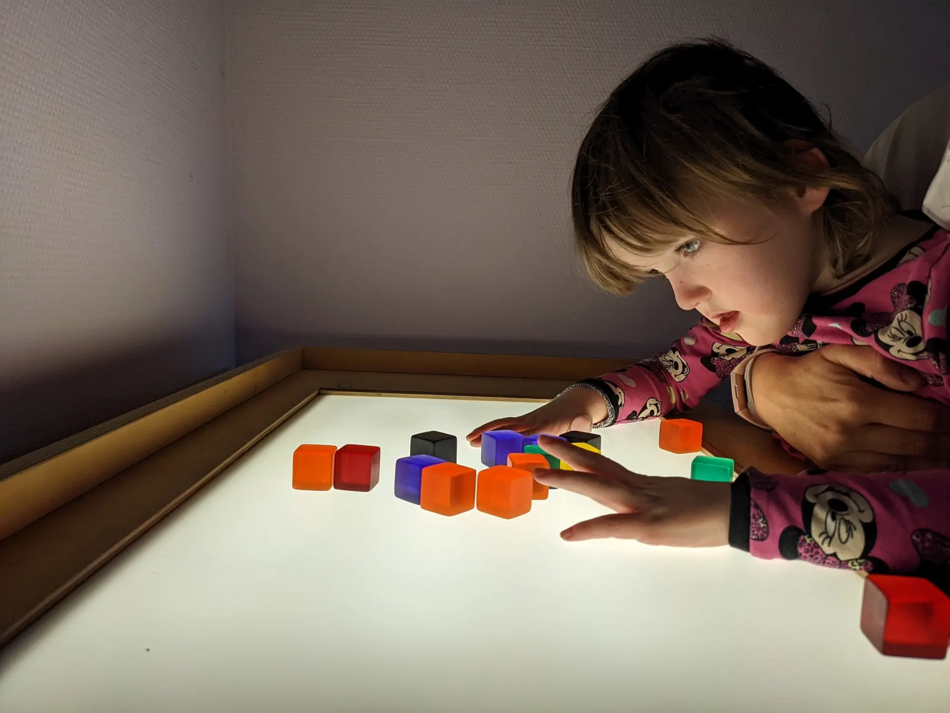 Ein Kind mit Komplexer Beeinträchtigung wird von einer pädagogischen Kraft ganz nah an einem Leichttisch gehalten. Dabei schaut es sich ganz nah bunte kleine Würfel, die auf der leuchtenden Oberfläche noch farbenfroher wirken. Mit einer Hand greift es zu einem der Würfel.