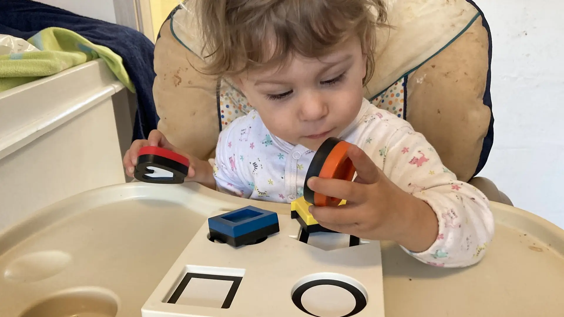 Ein kleines Kind sitzt im Kinderhochstuhl. Auf dem Stuhltablett vor ihm liegt ein Plastikpuzzle-Spiel mit vier Formen – Herz, Kreis, Quadrat und Haus. Dazu gibt es die gleichen Formen auf dem Spielbrett, damit das Kind sie korrekt darauf sortieren kann. Hier hält das Kind zwei der vier Formen in seinen Händen und schaut sich eine davon ganz nah und genau an.