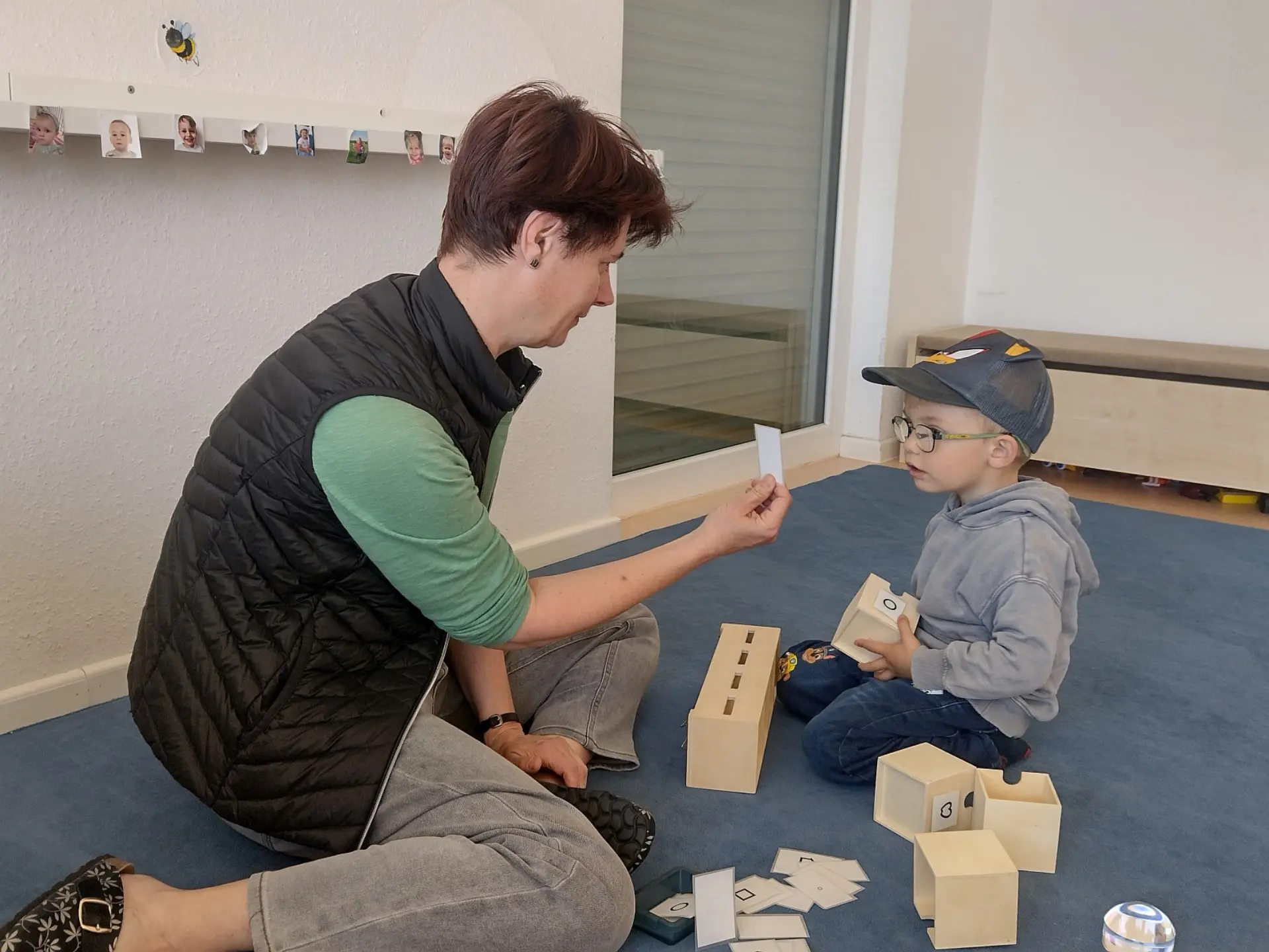 Eine Frühförderin und ein Kind sitzen auf einem Teppichboden in einem Kitaraum. Das Kind trägt eine Brille und schaut sich die Karte, die ihm die Sonderpädagogin zeigt. Auf dieser Karte und auf den restlichen 10-12 Kärtchen auf dem Boden sind Symbole wie Kreis oder Quadrat gezeichnet. Vor dem Kind steht eine Holzkiste mit vier Schlitzen auf dem Dach sowie vier Holzfächer mit jeweils einem der Symbole von den Kärtchen. Das Kind soll die Symbole erkennen und in das richtige Kastenschlitz hineinschieben.