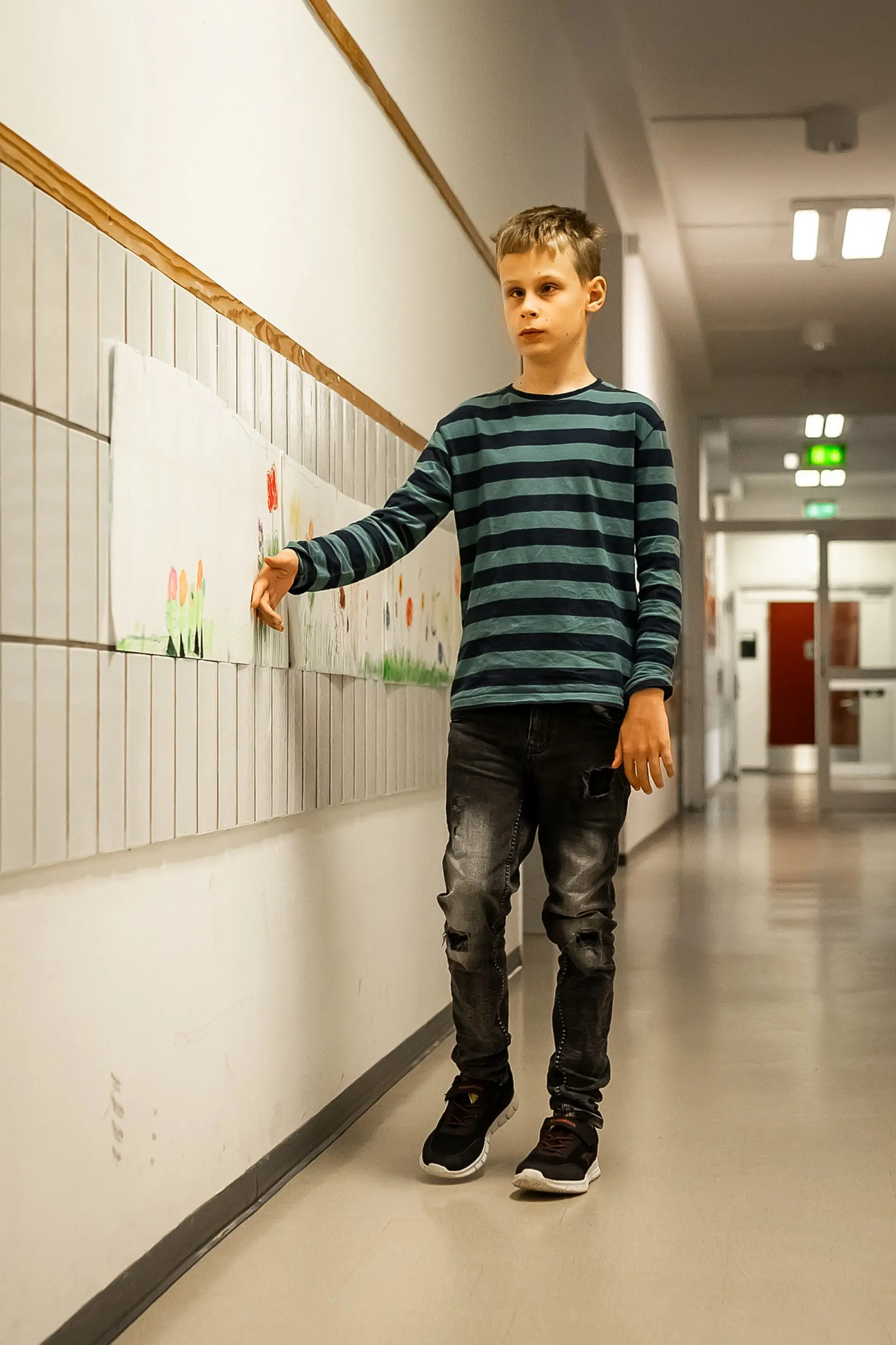 Ein Schüler mit Blindheit oder Sehbeeinträchtigung läuft im Gang des Schulgebäudes und ertastet mit dem Handrücken die Wand, damit er sich orientieren kann. An der Wand kleben Blätter mit von den Schüler*innen gemalten Bildern.