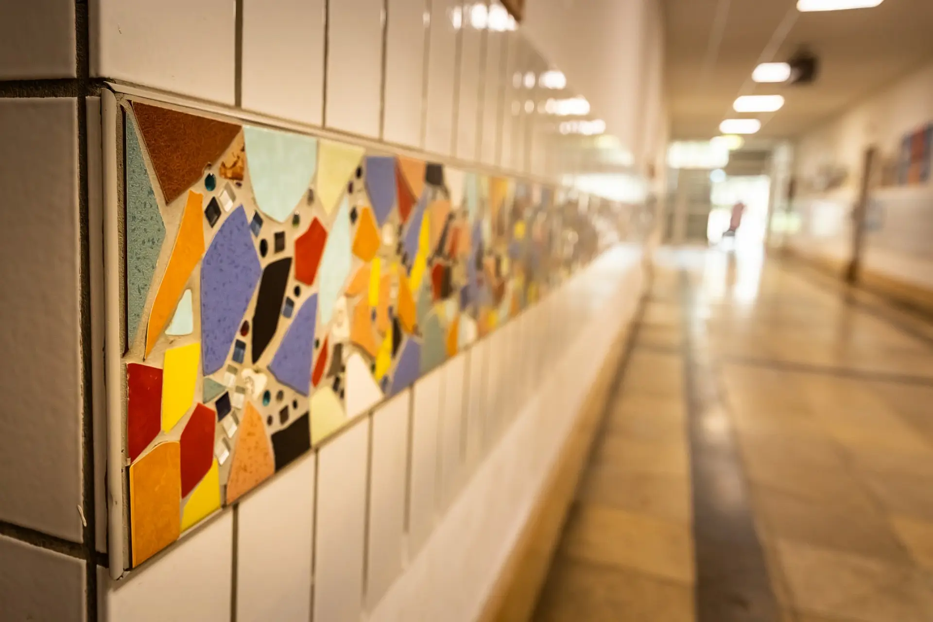 Ein langer Schulflur mit gefliesten Wänden. Auf Augenhöhe verläuft ein farbenfrohes Mosaikband, das aus bunten, unterschiedlich geformten Keramikstücken besteht. Die Fliesen reflektieren das Licht von oben. Der Flur führt in die Ferne, wo Türen und weitere Lichtquellen erkennbar sind.