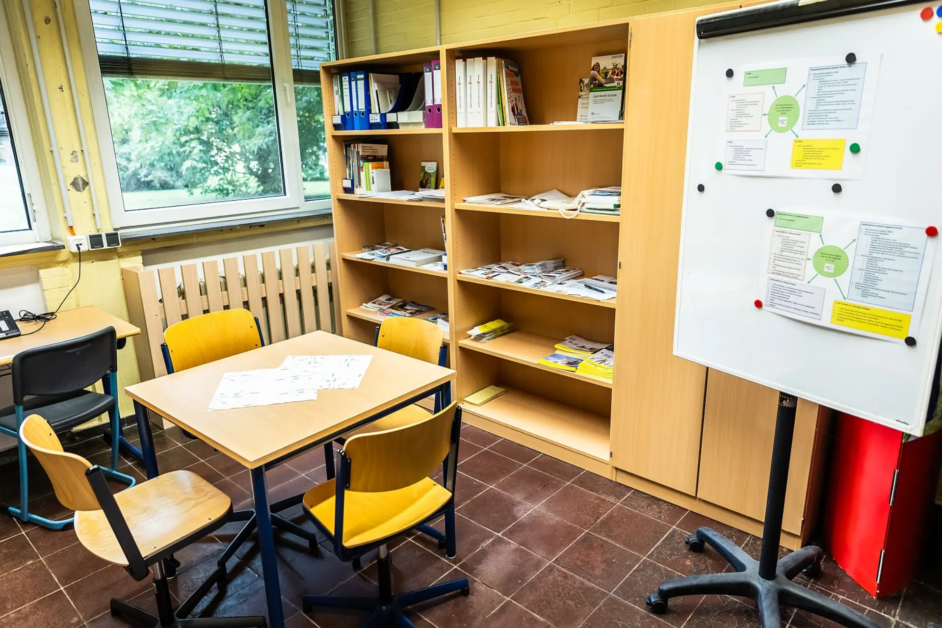 Ein Büroraum in einer Schule, der für die Berufsberatung der Schüler*innen genutzt wird. In der Mitte steht ein Holztisch mit vier Stühlen, bereit für die Gespräche. Dahinter befindet sich ein großes, offenes Holzregal, gefüllt mit Ordnern und Informationsmaterialien. Rechts daneben steht ein Whiteboard auf Rollen, an dem Informationsblätter mit Magneten befestigt sind. Links sind Fenster zu sehen, die Tageslicht hereinlassen und einen Blick ins Grüne ermöglichen. Der Raum hat einen Fliesenboden.