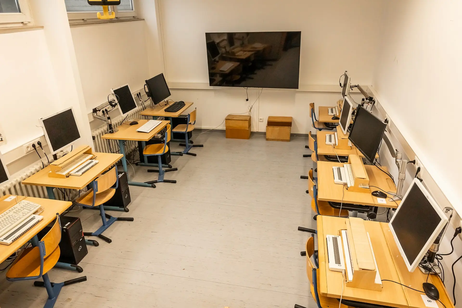 Ein aufgeräumter Computerraum im Souterrain der Schule. Man blickt in den Raum hinein, an den Wänden rechts und links ist jeweils eine Reihe von Holzschreibtischen aufgestellt. Die Tischen sind mit Computern, Monitoren und Tastaturen für Menschen mit Blindheit oder Sehbeeinträchtigung ausgestattet, vor jedem Tisch steht jeweils ein Stuhl. Ein großer Flachbildschirm hängt an der Wand im Hintergrund.