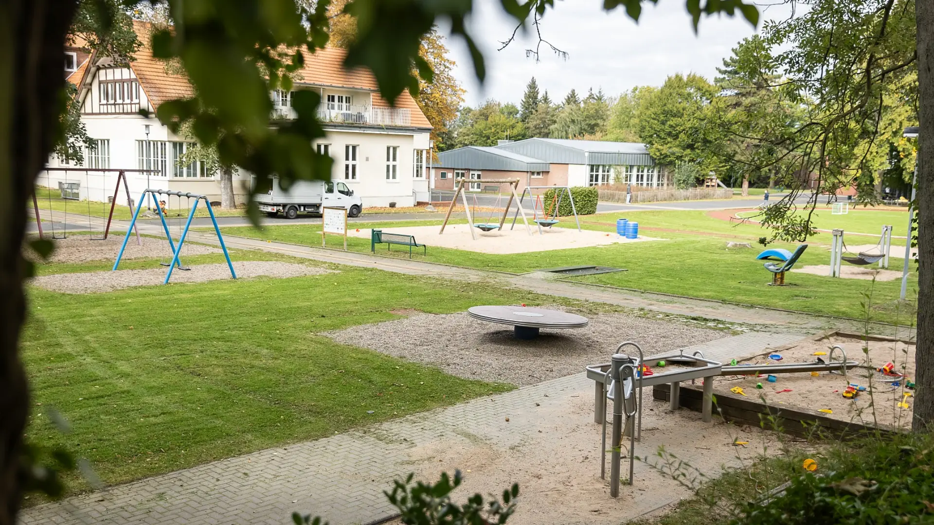 Ansicht eines Teils des Schulgeländes. Im Vordergrund ist ein großer Spielplatz mit verschiedenen Spielmöglichkeiten wie einem Sandkasten und einer Wasserspielanlage sowie vielen Schaukeln. Im Hintergrund ist ein Schulgebäude zu sehen.