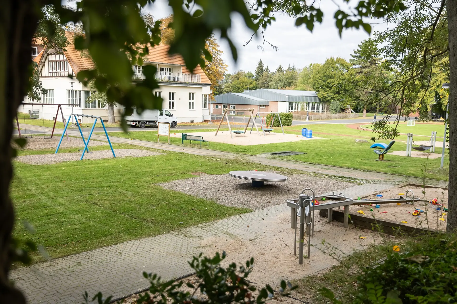 Ein Ausblick aufs Schulgelände zwischen den Bäume. Im Vordergrund ist ein großer Spielplatz mit verschiedenen Spielmöglichkeiten wie einem Sandkasten und einer Wasserspielanlage sowie vielen Schaukeln. Im Hintergrund ist ein Schulgebäude zu sehen.
