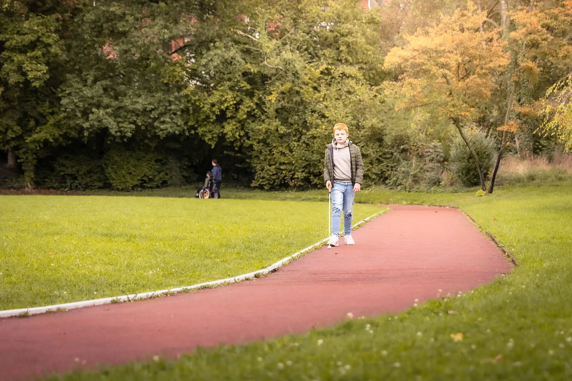 Ein Junge mit Blindenstock geht auf einem Spazierweg, im Hintergrund sieht man noch zwei Personen, die auch spazieren gehen – die eine Person sitzt im Rollstuhl, die andere schiebt diesen. Drum-herum sind noch Graswiesen und Bäume zu sehen.