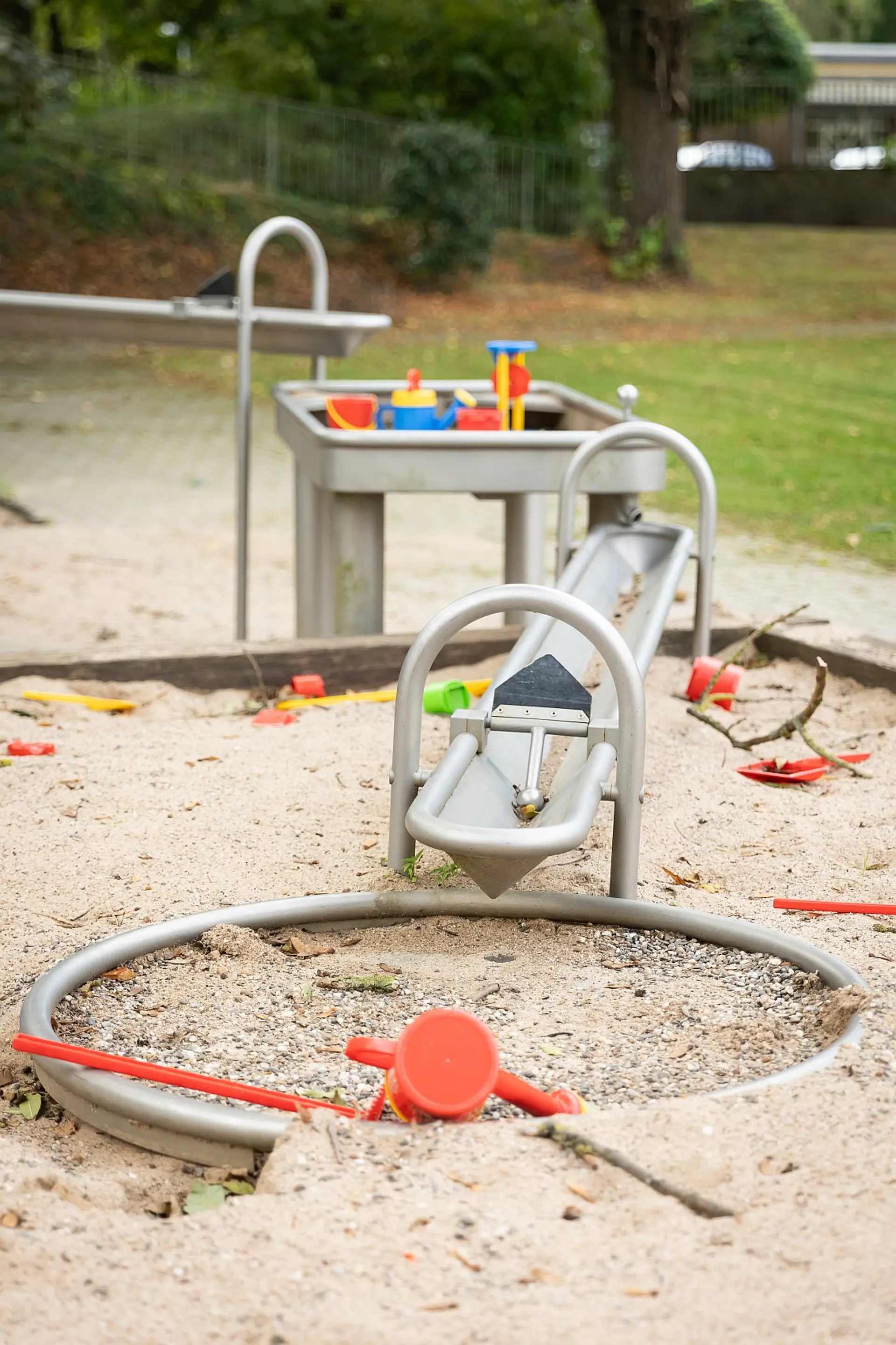 Ein Sandkastenbereich auf einem der Schul-Spielplätze mit verschiedenen Spielgeräten. Im Vordergrund befindet sich ein runder Metallrahmen im Sand, daneben liegen eine Plastik-Gießkanne und ein Stab. Dahinter ist eine Metallrampe mit einer Schaufel zu sehen, die in einen Sandhügel führt. Im Hintergrund steht ein Sand- und Wassertisch mit bunten Spielsachen. Das umliegende Gelände ist grün und baumbestanden.