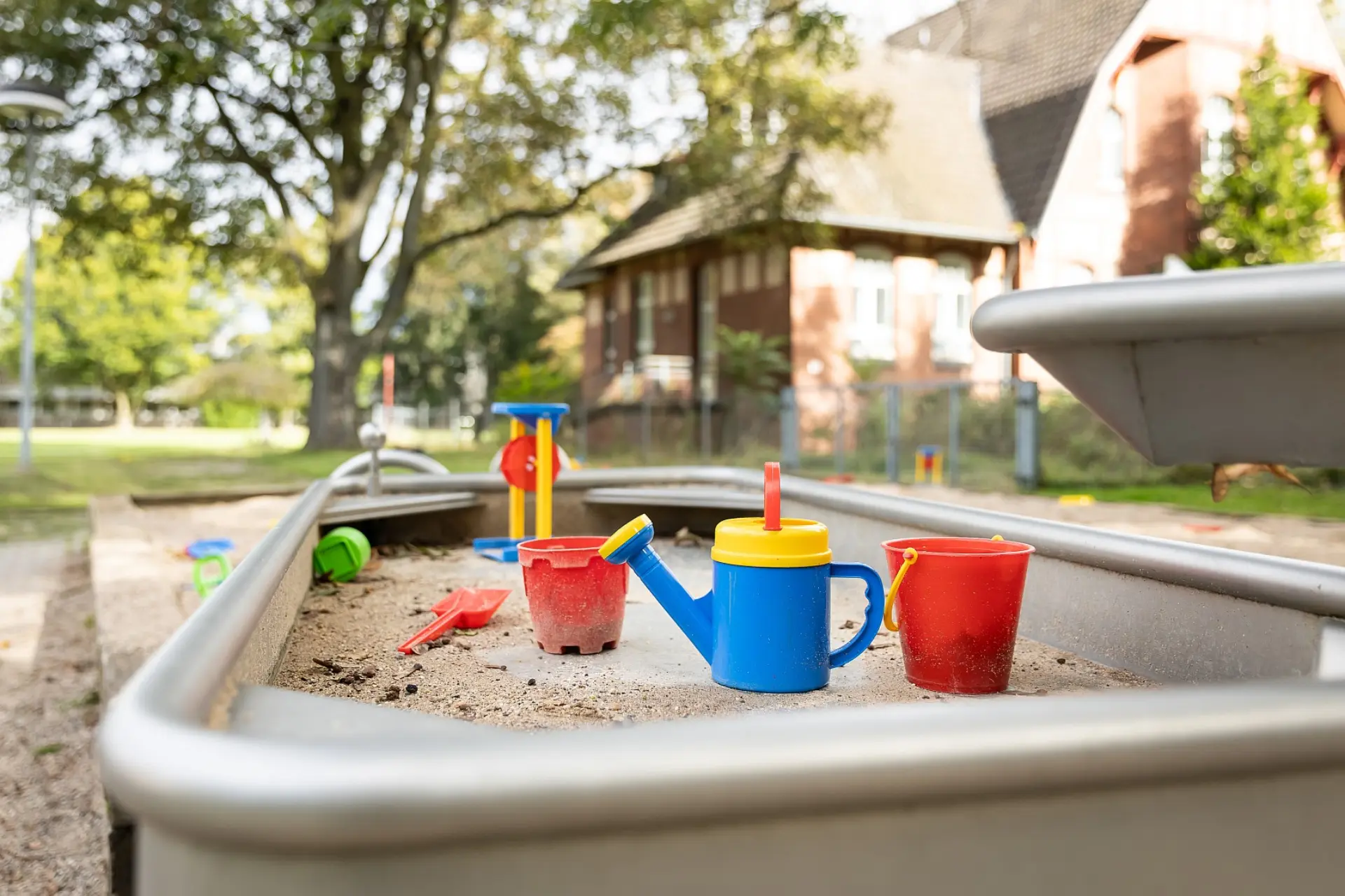 Eine Nahaufnahme eines Sand- und Wassertisches auf einem der Schul-Spielplätze. In dem mit Sand gefüllten Tisch stehen bunte Spielzeuge, darunter eine Gießkanne, rote Eimer, eine Schaufel und ein Ball. Im unscharfen Hintergrund sind Bäume und ein Gebäude zu sehen.