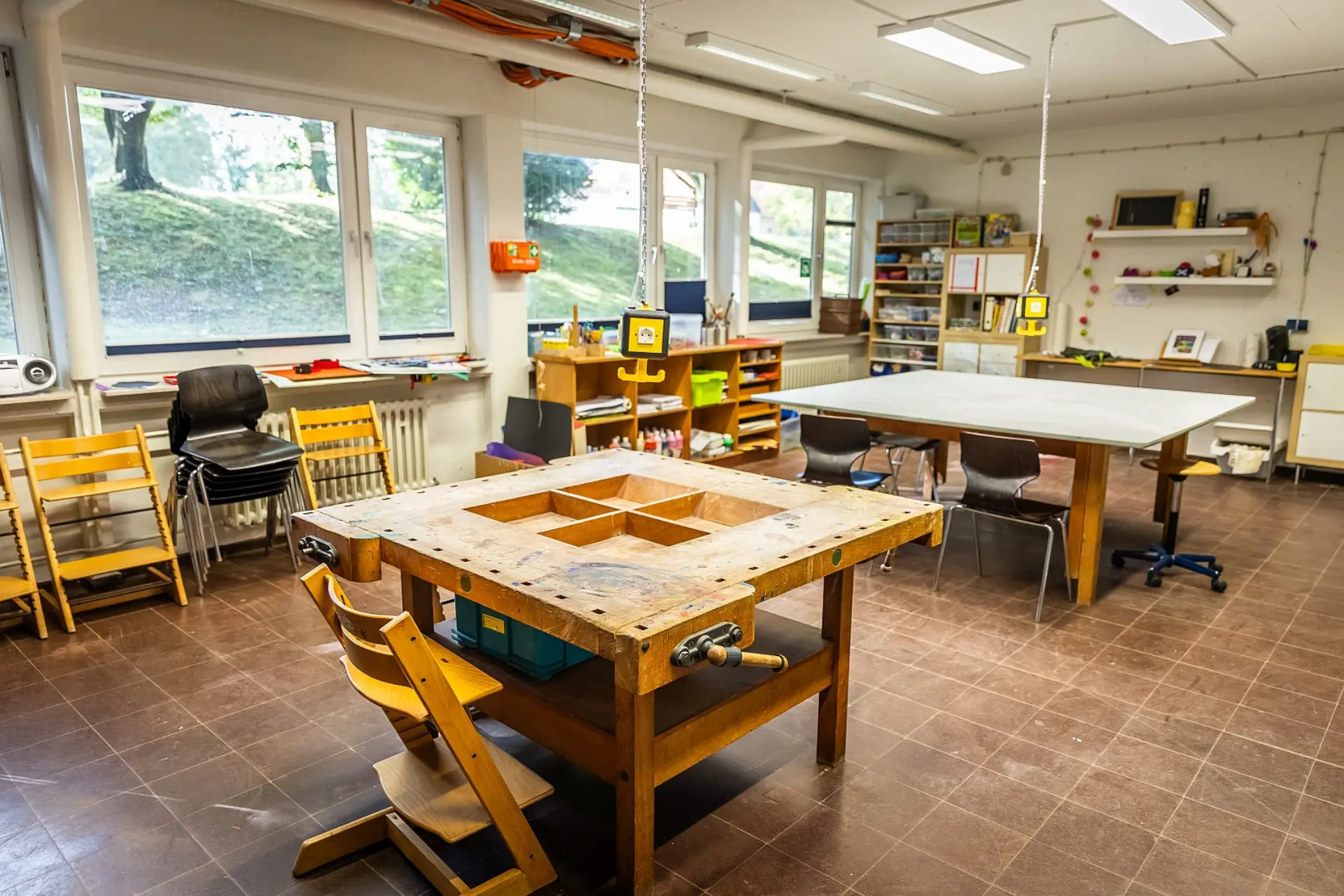 Im Vordergrund des Kunstraumes steht eine große, robuste Werkbank aus Holz mit eingeschnittenen Fächern und Schraubstöcken. Dahinter befinden sich andere Tische mit Stühlen, darunter ein großer Tisch für kreative Arbeiten. Entlang der Wände stehen Regale mit Mal- und Bastelmaterialien sowie Werkzeugen und weitere Stühle für die Schüler*innen. Große Fenster auf der linken Seite lassen viel Tageslicht herein und bieten einen Blick auf die Grünflächen im Schulhof. Der Raum hat einen Fliesenboden.