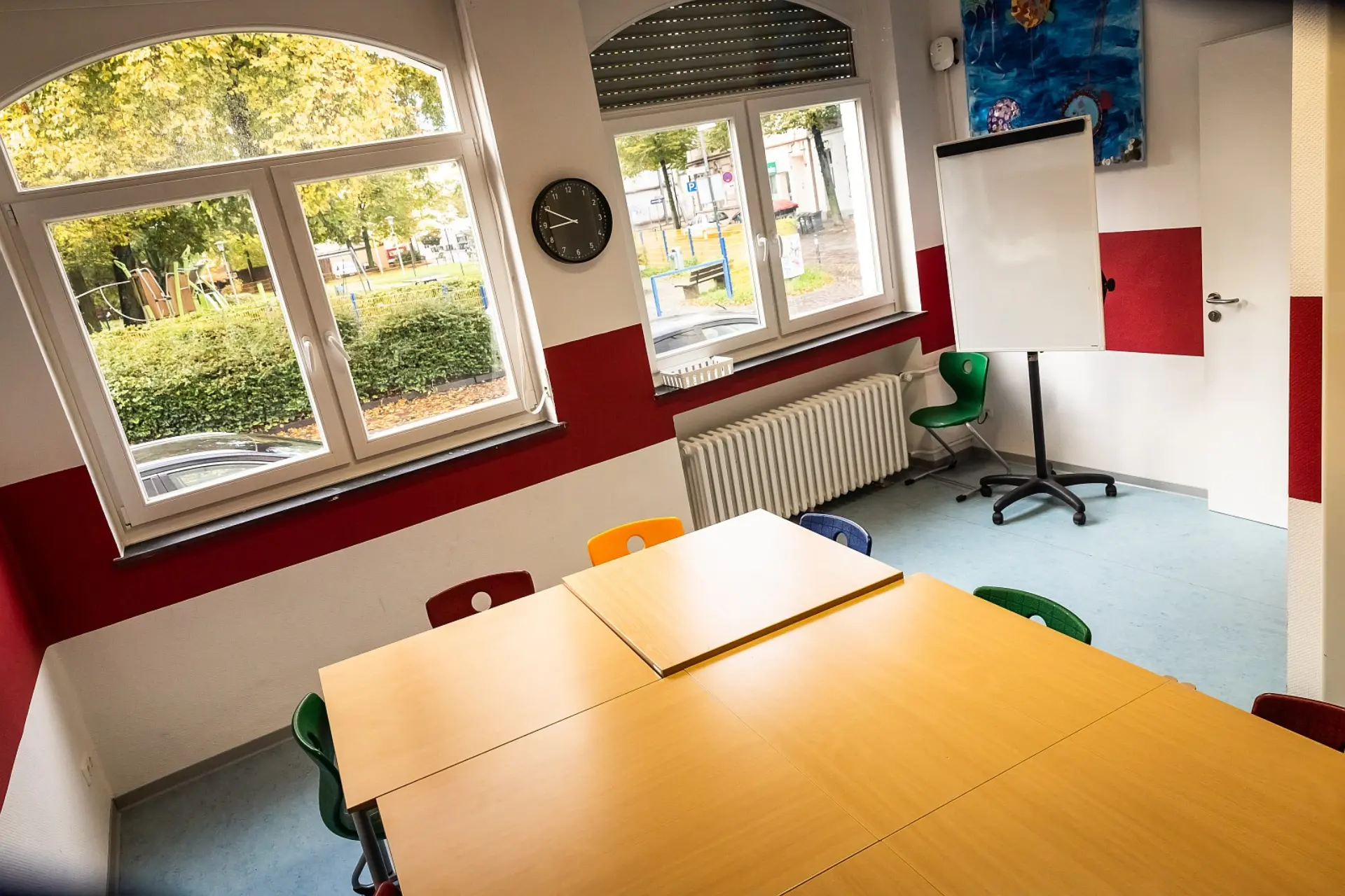 Ein heller Kursraum in der Schule, eingerichtet für Gruppenarbeiten oder Besprechungen. Sechs quadratische Tische sind so nebeneinander hingestellt, dass sie zusammen einen großen, viereckigen Holztisch bilden. Dieser steht im Vordergrund, umgeben von Stühlen in verschiedenen Farben. Die Wände sind zweifarbig gestrichen. Große Fenster auf der linken Seite bieten einen Blick auf Bäume und einen Spielplatz des Schulhofs. Rechts im Raum befindet sich ein Whiteboard auf Rollen und eine Tür.