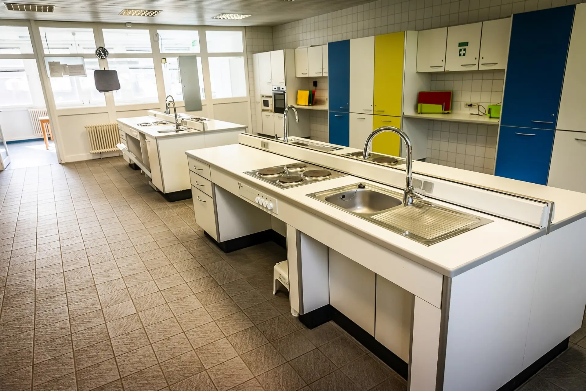 Ein weitläufiger Blick auf eine helle Lehrküche in einer Schule. Zwei große Kochinseln stehen parallel zueinander auf einem Fliesenboden. Jede Insel ist mit mehreren Herdplatten und Spülbecken ausgestattet. Im Hintergrund erstreckt sich entlang der Wand eine Reihe von Küchenschränken, die eine funktionale Atmosphäre schaffen. Große Fenster auf der linken Seite lassen viel Tageslicht herein.