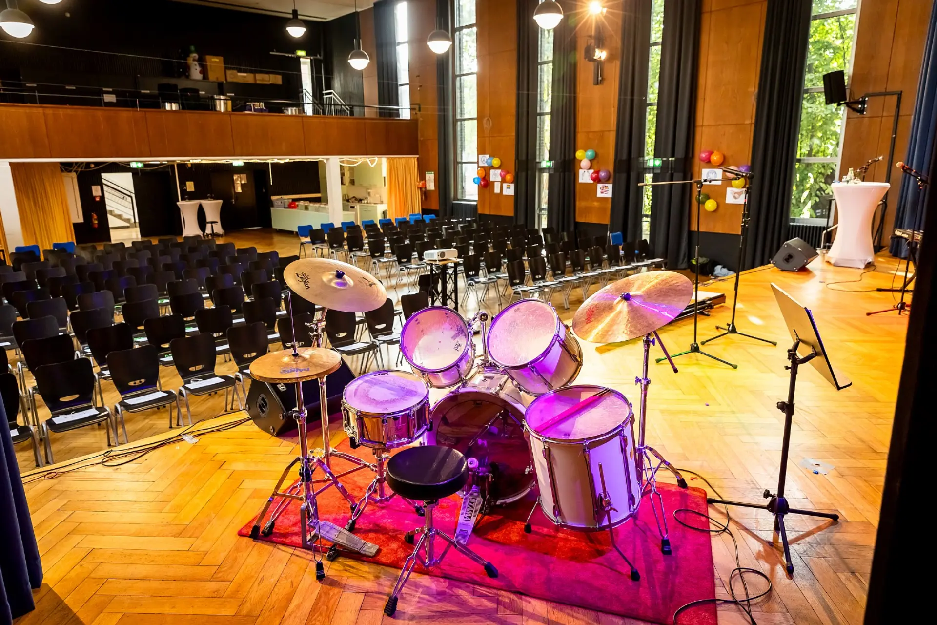 Ein Schlagzeug-Set steht auf der Bühne mit Holzboden in der schulischen Aula. Dahinter sind Reihen von leeren Stühlen zu sehen, die auf die Bühne ausgerichtet sind. Links oben befindet sich ein Balkon mit weiteren Sitzplätzen. Im Hintergrund sind hohe Fenster mit schweren Vorhängen, Holzwände und verschiedene Veranstaltungsausstattungen wie Stehtische und Ballons zu erkennen.