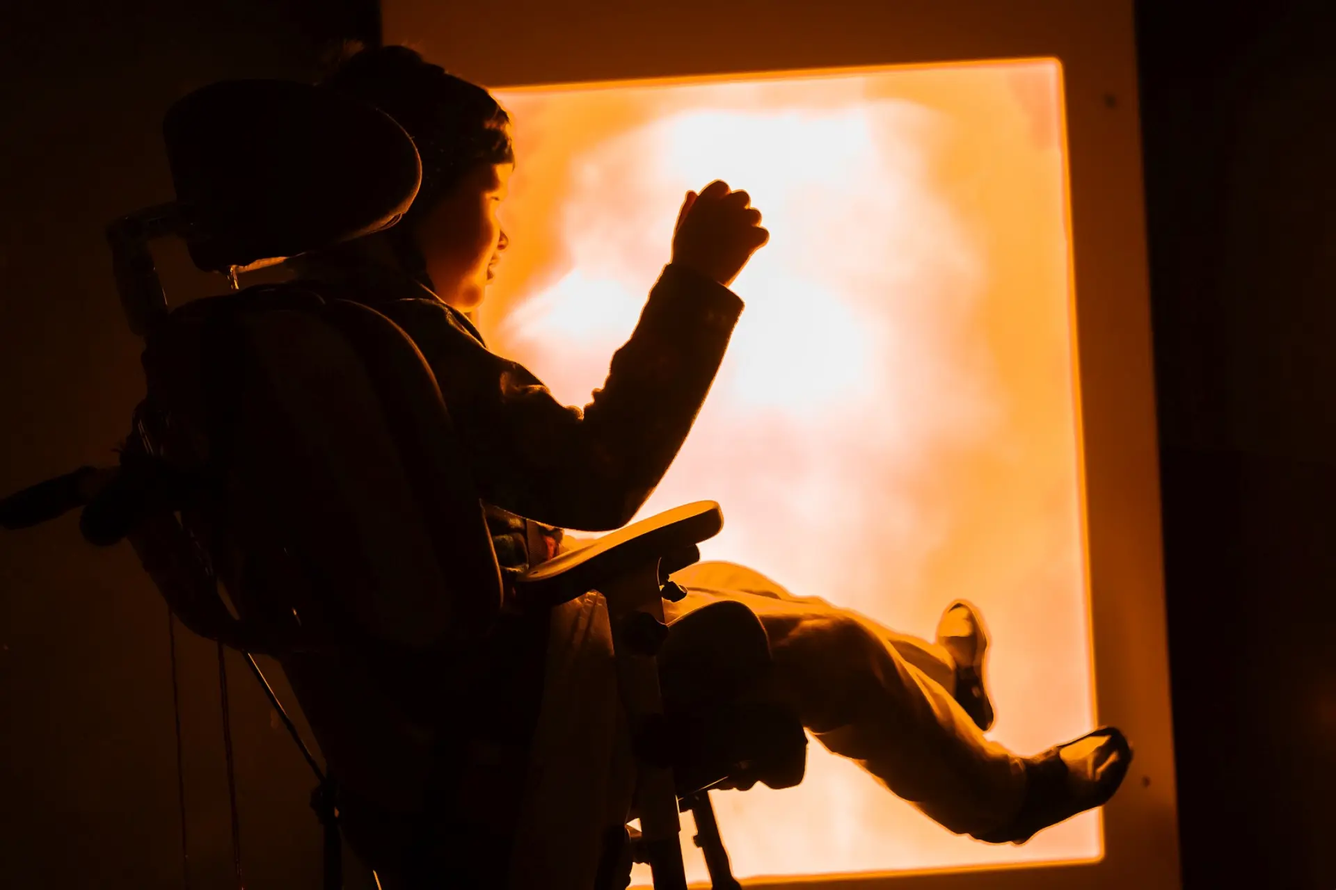 Ein Schüler mit komplexer Beeinträchtigung sitzt in seinem Rollstuhl in einem komplett abgedunkelten Raum. Links von ist eine eingeschaltete körpergroße Lichtplatte. Er schaut zu dieser Lichtfläche und hebt den einen Arm in ihre Richtung.