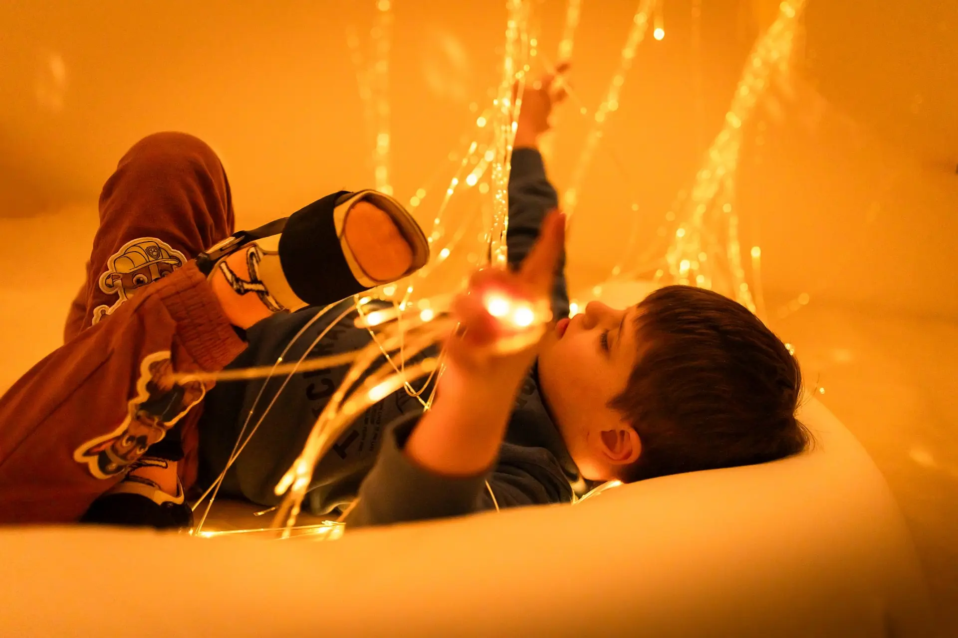 Ein kleiner Junge mit komplexer Beeinträchtigung liegt auf einer Matratze in einem abgetrennten Raum, im so genannten Snoezelraum zum Entspannen. Über ihn hängt eine feine Lichterkette mit vielen Strängen. Manche davon liegen auf ihm und er greift mit seinen Händen zu ihnen, indem er sie sich ganz genau anschaut.