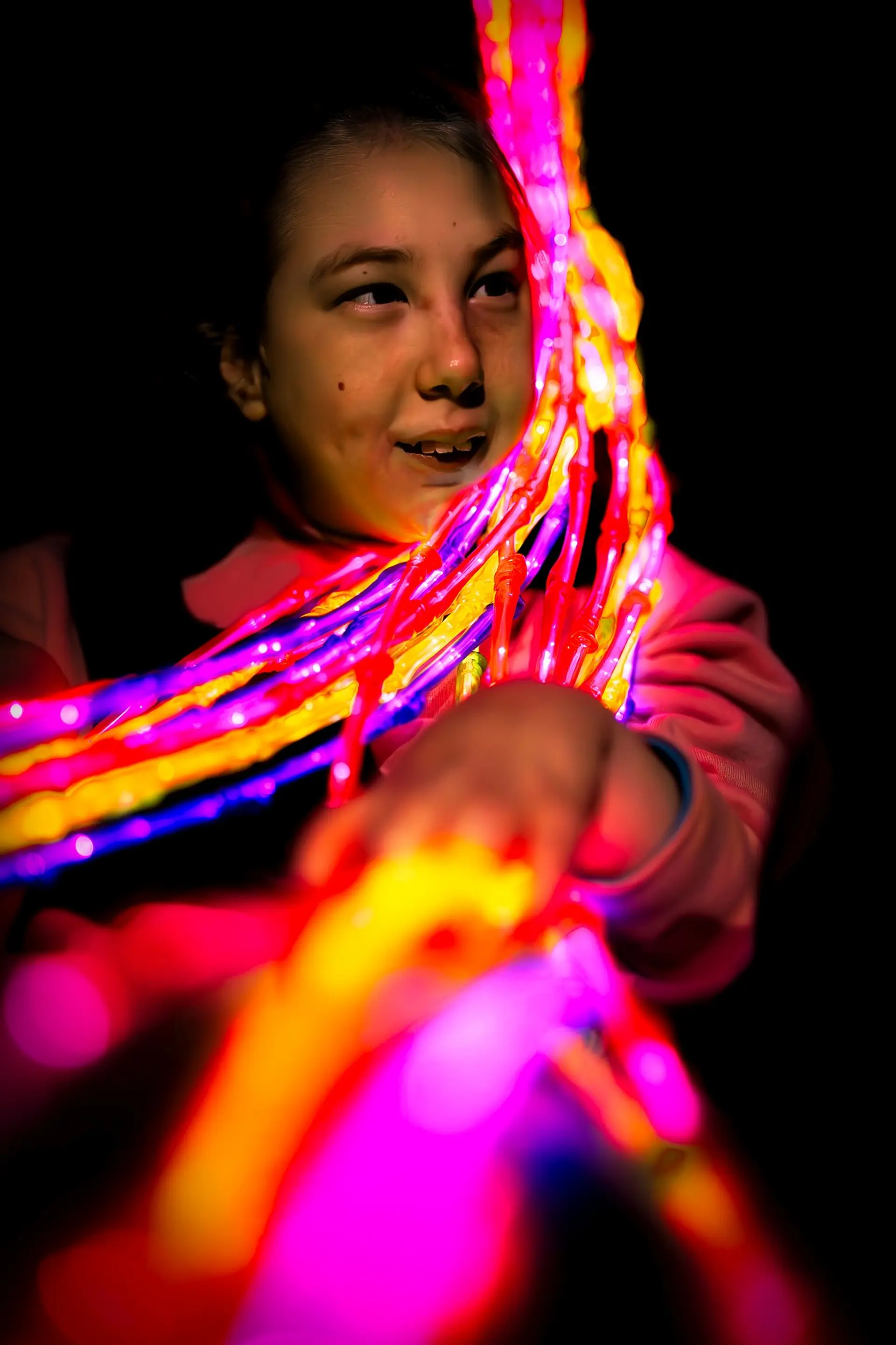 Schülerin mit komplexer Beeinträchtigung in einem komplett abgedunkelten Raum, dem so genannten "Low-Vision-Raum" der Schule. Über über Schulter hängt eine Lichterkette mit vielen Strängen und in verschiedenen Farben. Sie schaut zum bunten Licht und streckt ihren Arm, um die Kette zu berühren. 