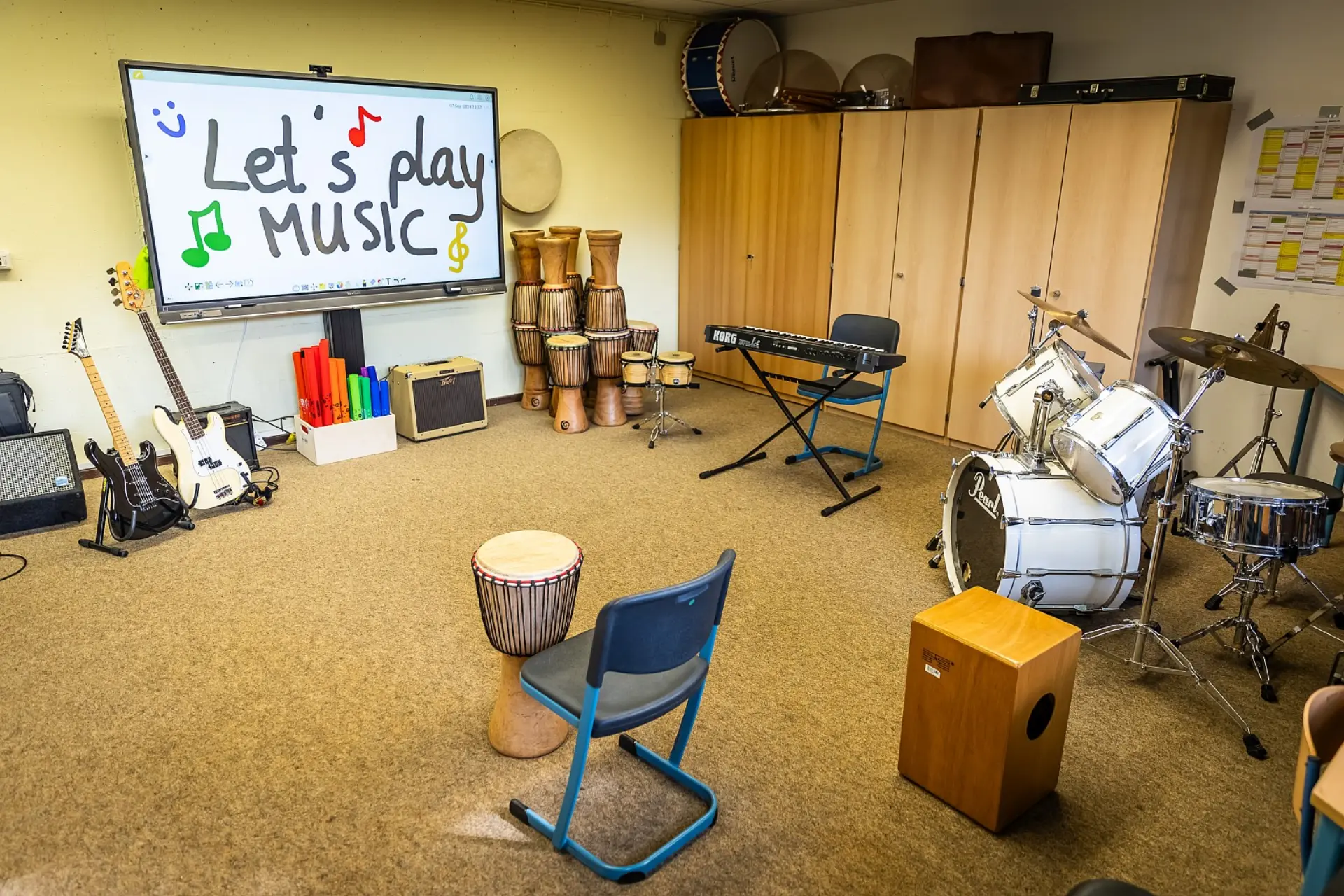 Ein Musikraum, der mit verschiedenen Instrumenten ausgestattet ist. Ein großes, interaktives Whiteboard an der Wand zeigt die Aufschrift "Let's play music!". Im Raum verteilt stehen eine E-Gitarre, verschiedene Trommeln (Djemben und ein Schlagzeug), ein Keyboard und eine Cajon. Im Hintergrund befinden sich Holzschränke und an der Wand hängt ein Jahreskalender aus Papier. Der Raum hat einen Teppichboden.