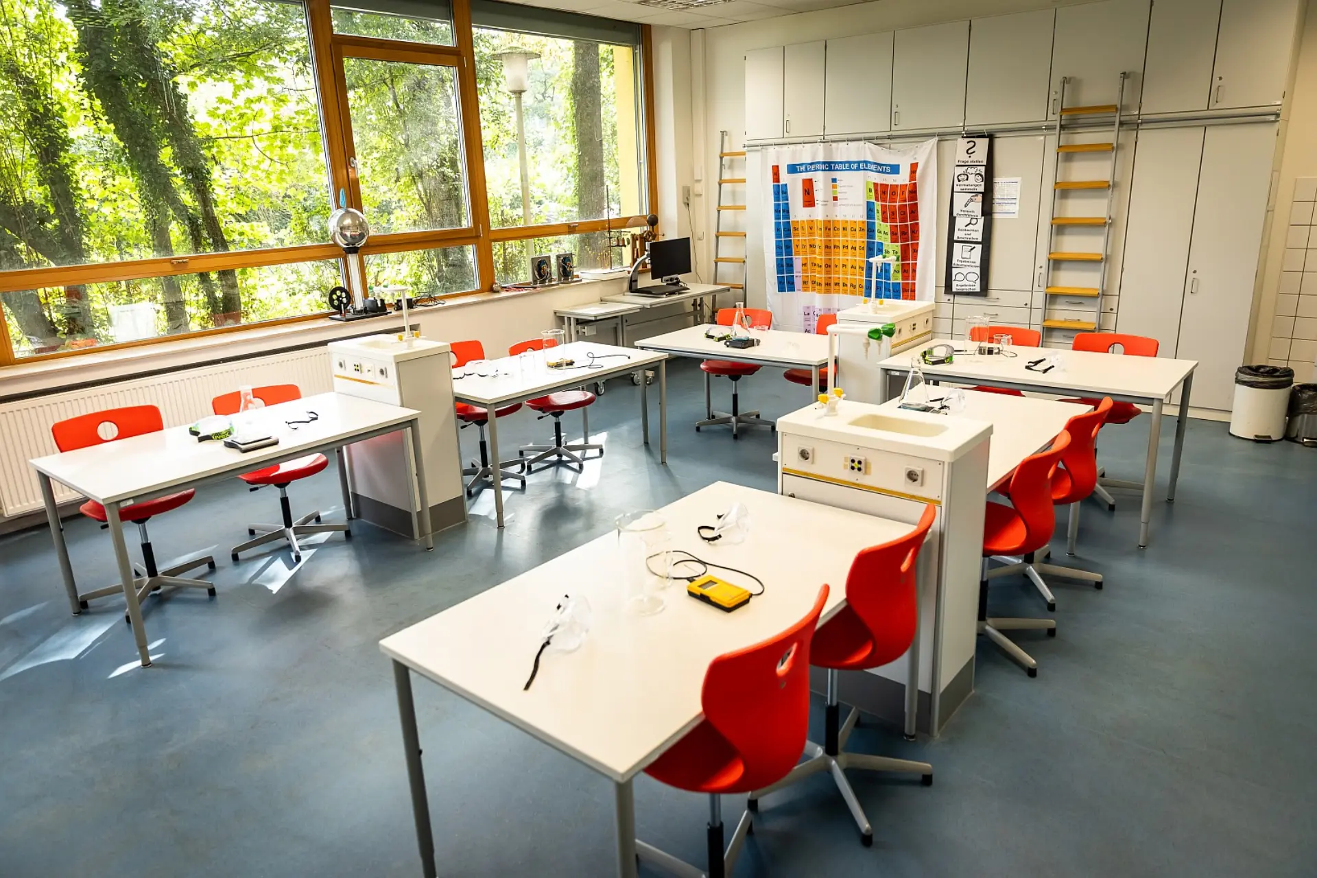 Im geräumiger Chemie- und Physikraum sind mehrere Labortische mit Stühlen verteilt. An drei Stellen neben den Tischen stehen integrierte Spülbecken und Wasserhähne. Die linke Wand ist mit großen Fenstern versehen, die einen Blick auf die Bäume im Schulhof ermöglichen. An der Wand uns gegenüber befinden sich viele hohe Schränke sowie zwei Leiter, damit man die oberen Schränke erreichen kann. An einer Leiste von den Schränken hängt ein Stoffplakat mit dem Periodensystem der Elemente. Verschiedene Laborgeräte wie sprechende Thermometer, Kolben und Schutzbrillen sind auf den Tischen bereitgestellt.