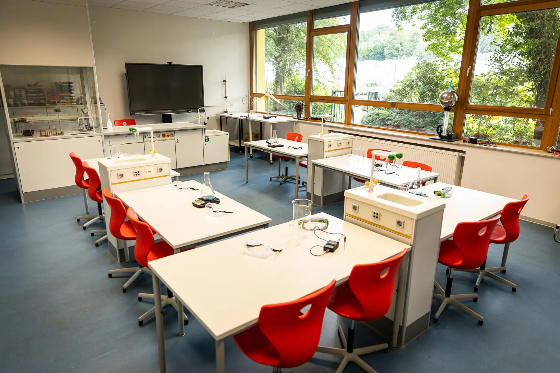 Mehrere Labortische mit integrierten Spülbecken und Wasserhähnen sind im Fachraum verteilt, umgeben von leuchtend roten Stühlen. Jeder Tisch ist mit verschiedenen Laborgeräten und Utensilien für Experimente ausgestattet. Im Hintergrund befindet sich ein großer Flachbildschirm an der Wand und eine Vitrine mit weiteren Geräten. Große Fensterfronten lassen viel Tageslicht herein und bieten einen Blick auf Bäume.