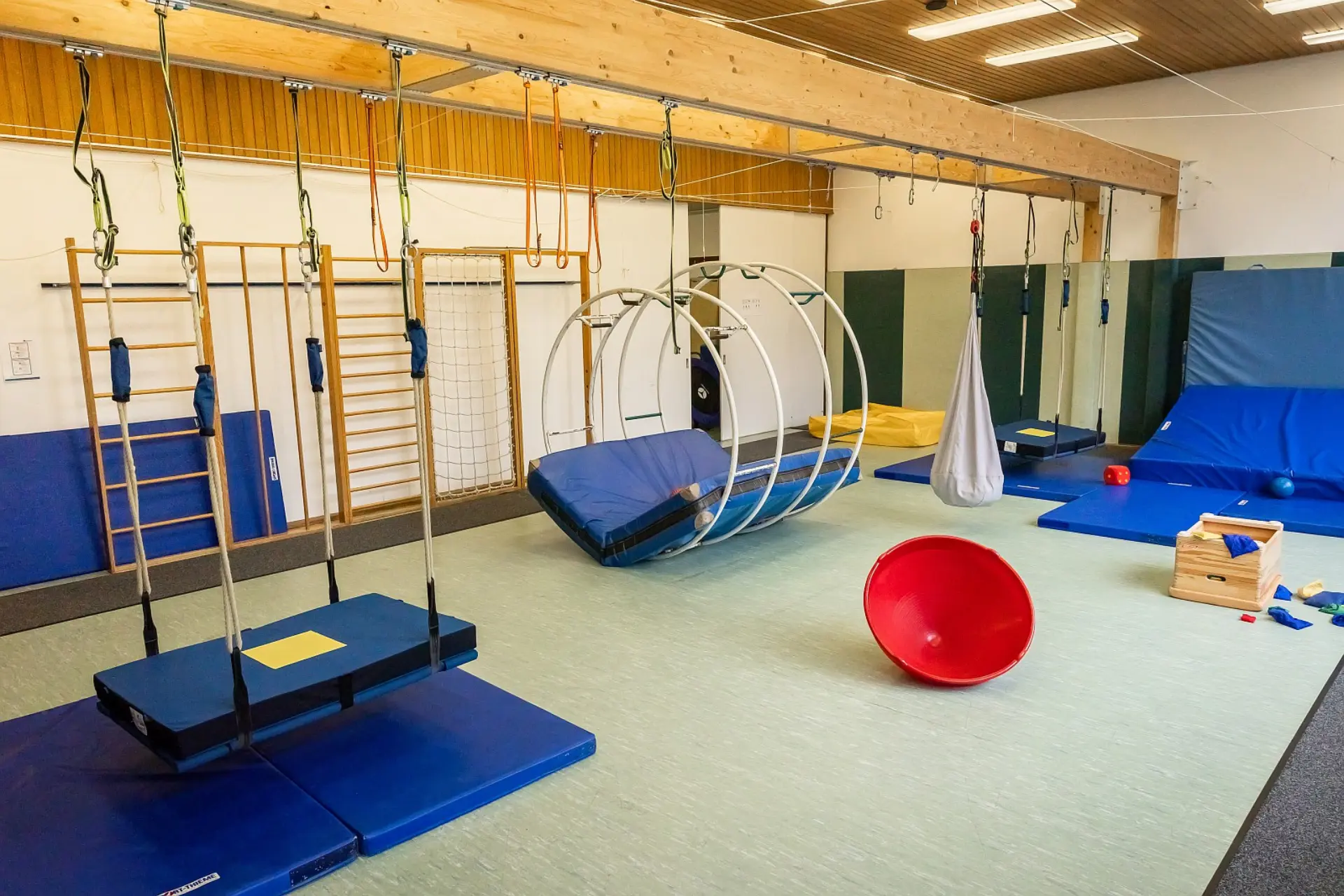 Ein großzügiger Gymnastikraum in der Schule, der speziell für Schüler*innen mit komplexen Beeinträchtigungen ausgestattet ist. Im Raum befinden sich verschiedene therapeutische und sportliche Geräte: eine Schaukelplattform, Sprossenwände, ein großes Rad mit einer Matte und ein Hängesack. Der Boden ist mit mehreren Matten ausgelegt, die Sicherheit bieten. Verschiedene kleine Spiel- und Therapiegeräte, wie ein Halbrund und Holzkisten mit Bauklötzen, sind ebenfalls sichtbar.