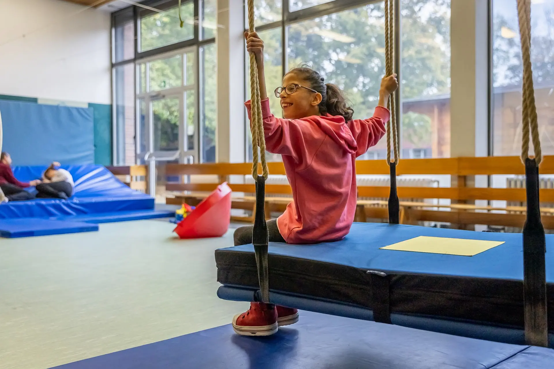 Eine Schülerin mit körperlicher Beeinträchtigung sitzt auf einer der Schaukel in der Gymnastikhalle, hält sich mit zwei Händen für die Schaukelseile und lächelt. Im Hintergrund sieht man noch zwei Personen, die auf dicken Matten auf dem Boden liegen, sowie andere Spielgeräte. Die eine Wand der Halle ist komplett mit Fenstern, sodass der Raum sehr hell ist. Vor den Heizungskörpern vor der Fensterfront ist eine Holzbrettkonstruktion aufgebaut, damit sich die Schüler*innen nicht verletzen.
