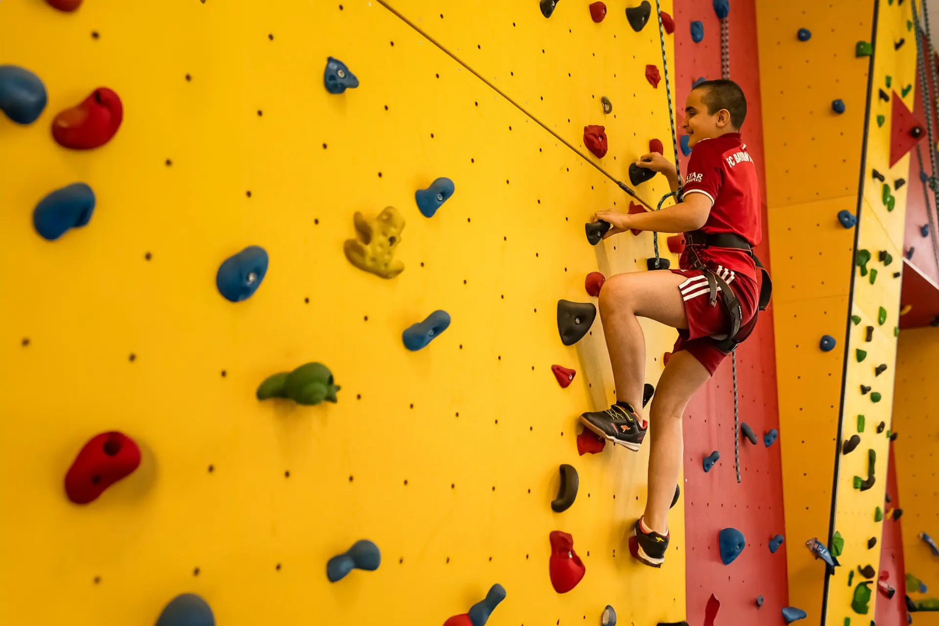 Ein Schüler mit Sehbeeinträchtigung klettert an einer Kletterwand. die mit vielen Klettergriffen in verschiedenen Farben und Formen ausgestattet ist. Der Schüler trägt Sportkleidung und wird mit einem Seil von oben gesichert. 