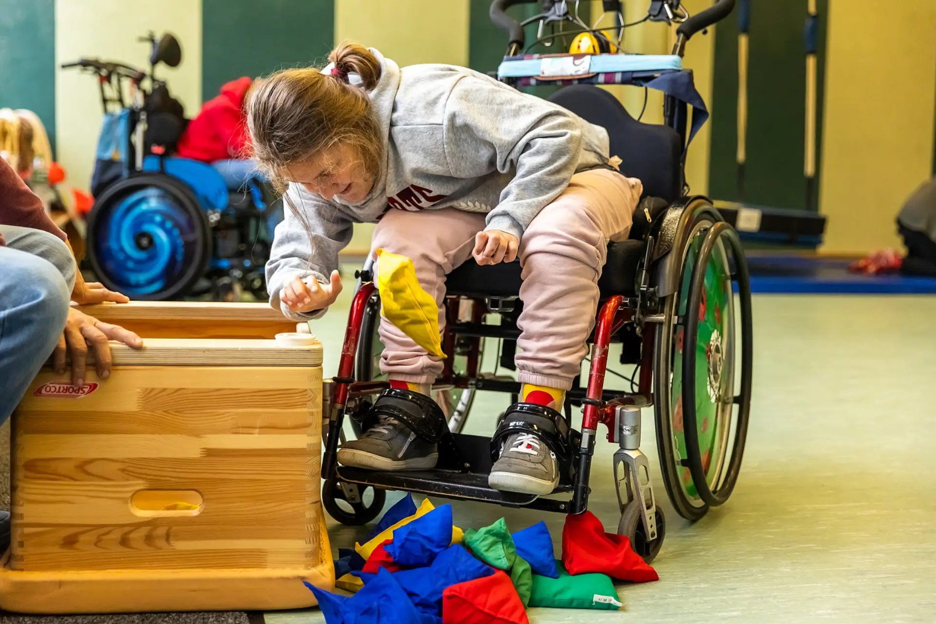 Eine Schülerin im Rollstuhl beugt sich über einer Holzkiste und nimmt mit der einen Hand handgroße Stoffsäckchen voller Sand als Spiel heraus. Sie befindet sich in der Therapiehalle der Schule, im Hintergrund sieht man noch eine junge Person im Rollstuhl sowie Schutzmatten und Geräte.