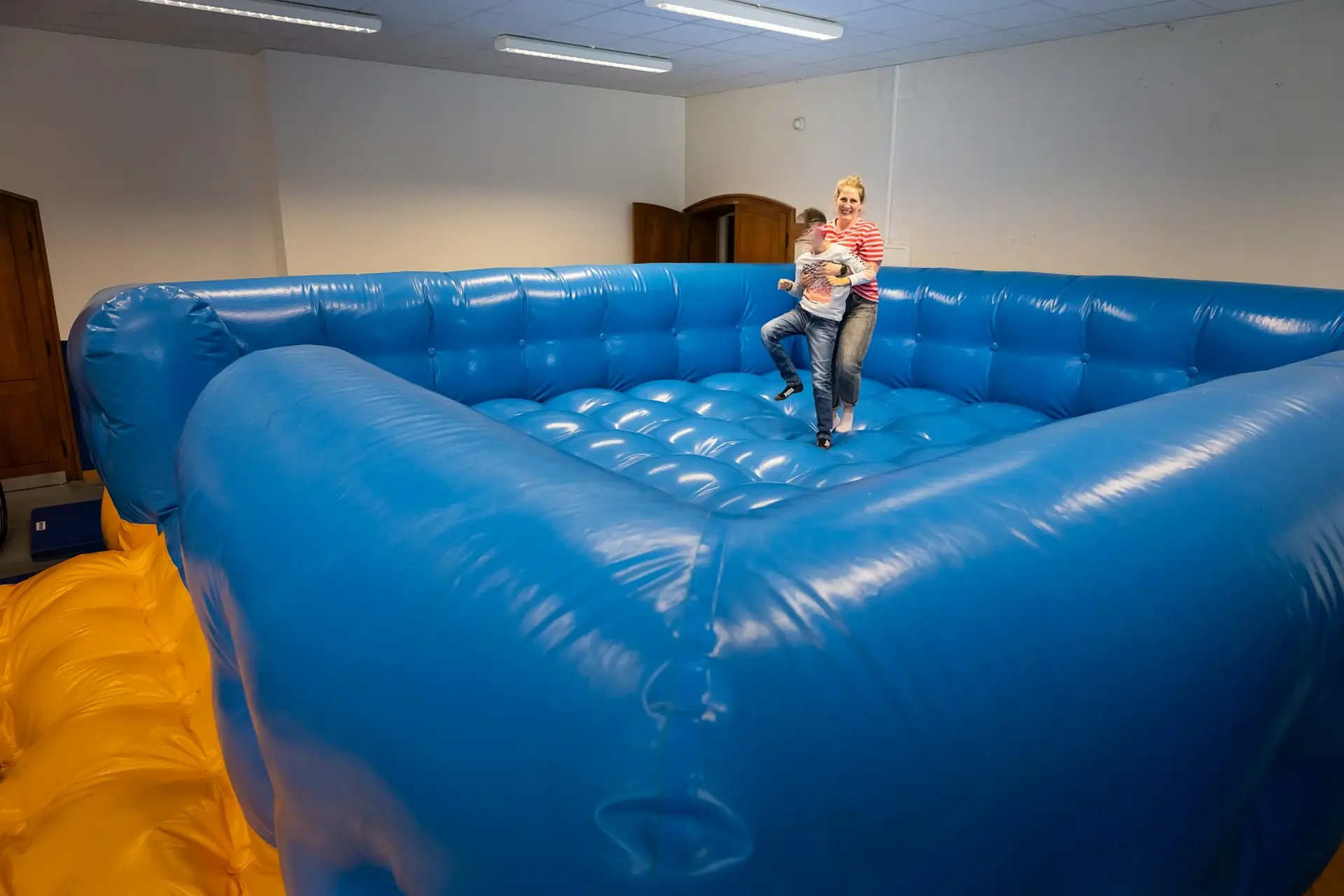 Ein sehr großer, aufgeblasener Airtramp mit dicken Wänden erfüllt einen Raum in der Schule. In seiner Mitte befinden sich eine Pflegerin und ein Schüler mit körperlicher Beeinträchtigung. Sie hält ihn von hinten, damit er aufrecht stehen und mit den Füßen trampeln kann.