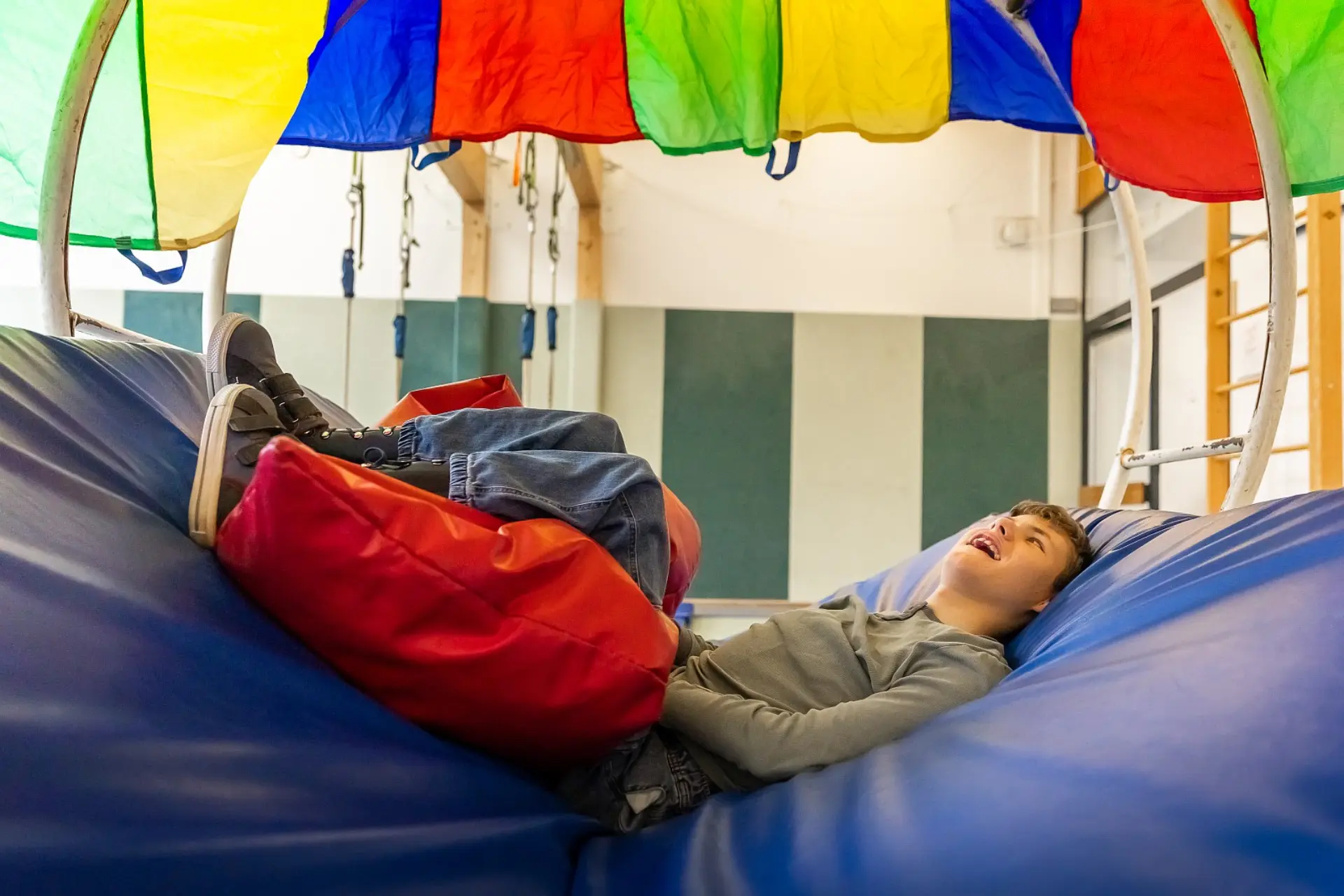 Ein Schüler mit körperlicher Beeinträchtigung liegt in einer Rundenschaukel in der Gymnastikhalle. Unter seinem Körper sind Sportmatten und unter seinen Beinen dicke Kissen ausgelegt, damit er bequem liegt, über der Schaukel ist eine große bunte Stoffplane. Er schaut zu ihr und scheint sich darüber zu freuen.