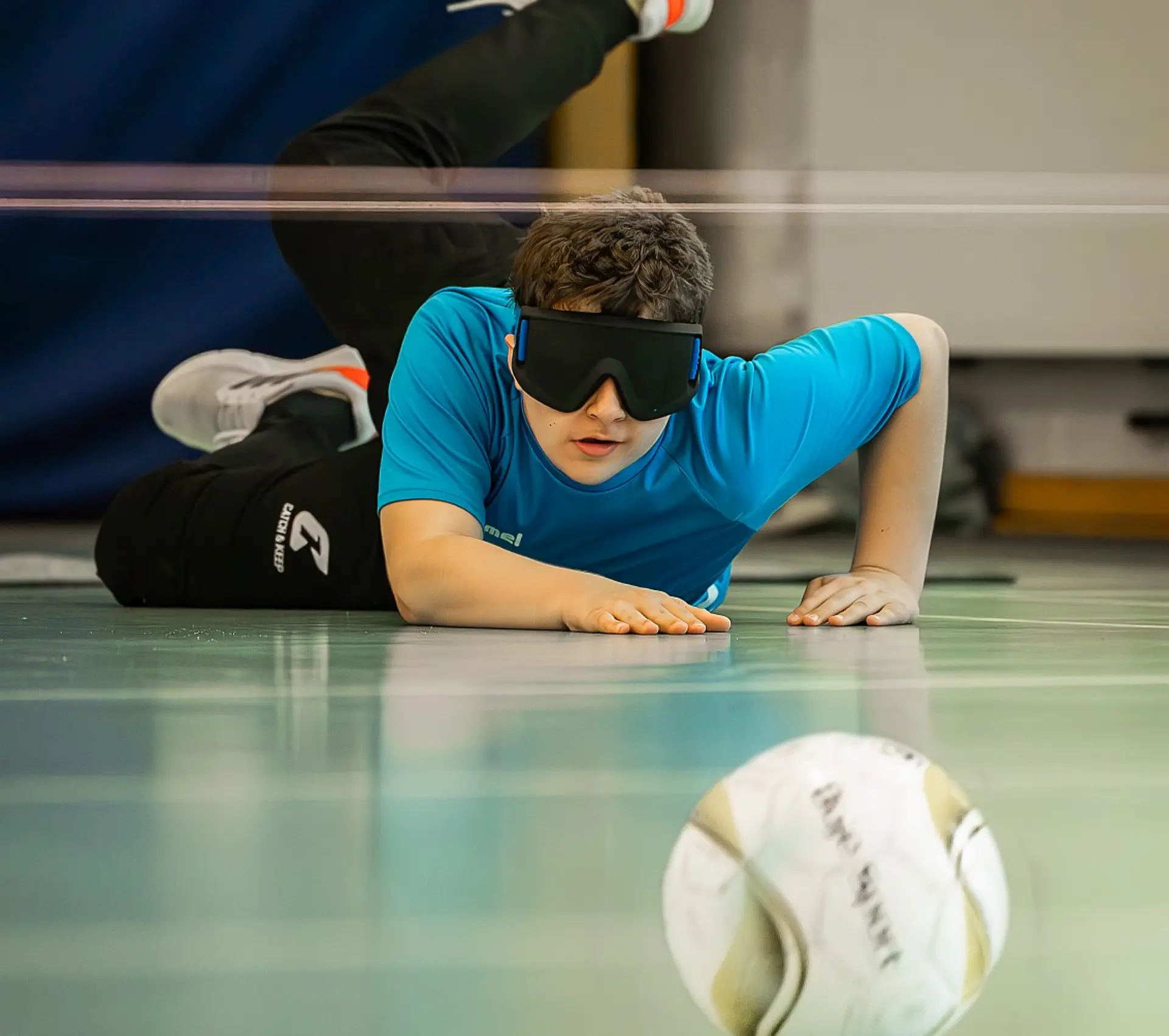 Ein Schüler in der Sekundarstufe I spielt Torball in der Sporthalle der Schule. Er trägt eine Dunkelbrille, liegt auf dem Bauch auf dem Boden und stützt sich mit beiden Händen, sodass sein Oberkörper nach oben ragt. Er sieht so aus, als ob er gerade den Ball geworfen hat und dabei nach vorne und zum Boden gesprungen ist. Im Vordergrund sieht man den hüpfenden Ball.