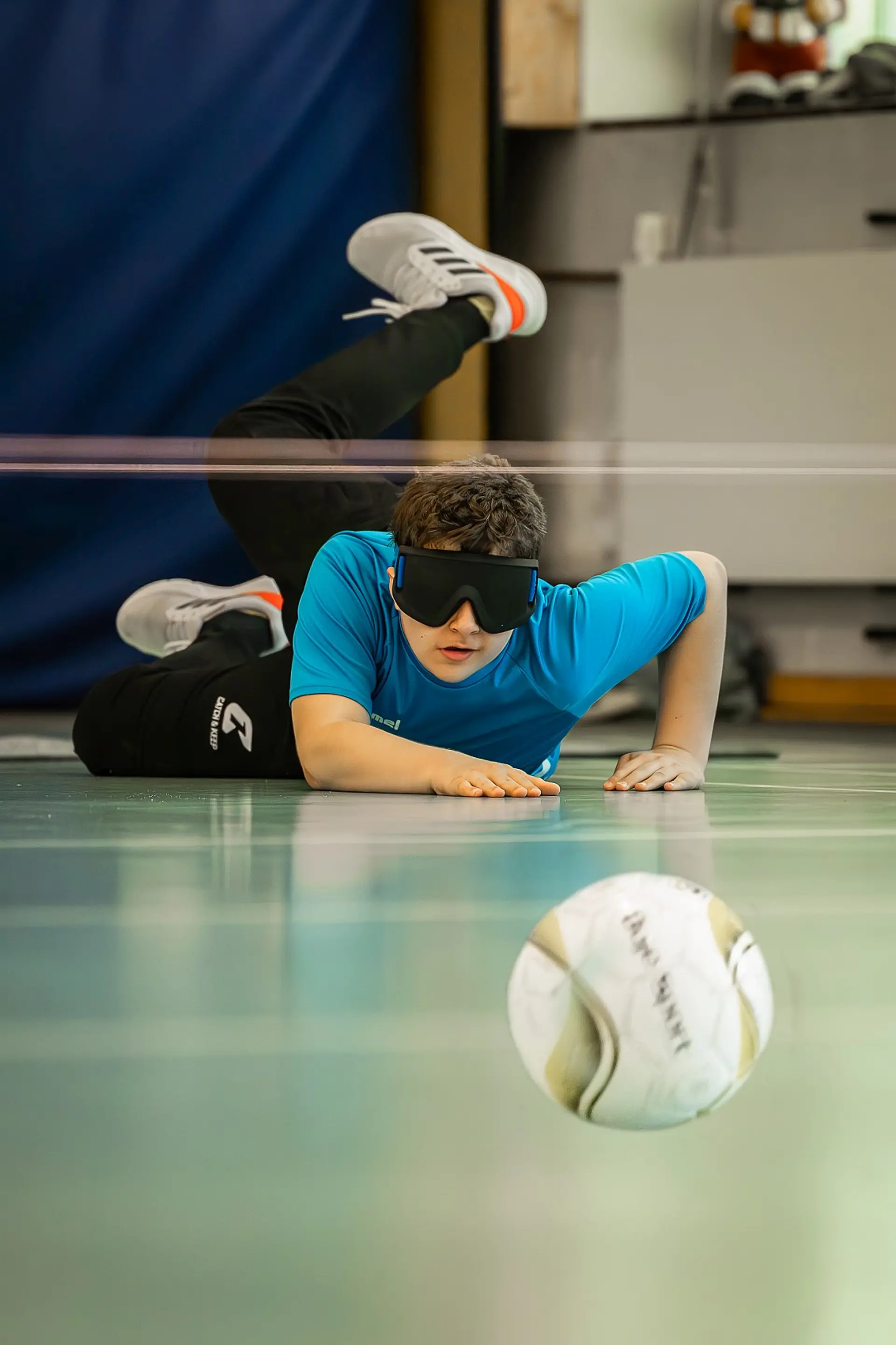 Ein Schüler in der Sekundarstufe I spielt Torball in der Sporthalle der Schule. Er trägt eine Dunkelbrille, liegt auf dem Bauch auf dem Boden und stützt sich mit beiden Händen, sodass sein Oberkörper nach oben ragt. Er sieht so aus, als ob er gerade den Ball geworfen hat und dabei nach vorne und zum Boden gesprungen ist. Im Vordergrund sieht man den hüpfenden Ball.