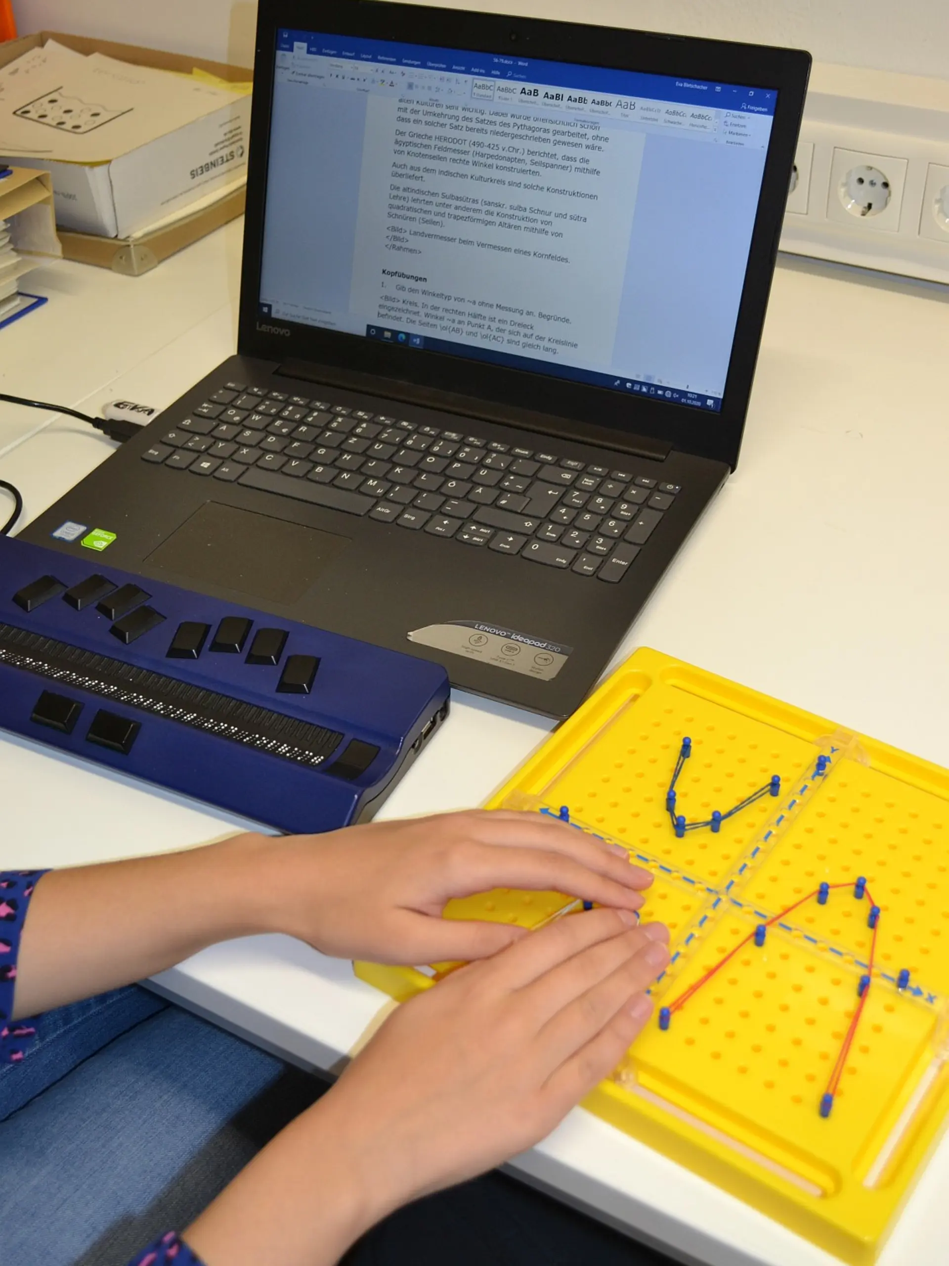 Die Hände einer Schülerin, die an ihrem Laptoparbeitsplatz mit Braillezeile sitzt und auf dem danebenliegenden Koordinatensteckbrett einen Graphen abtastet. 