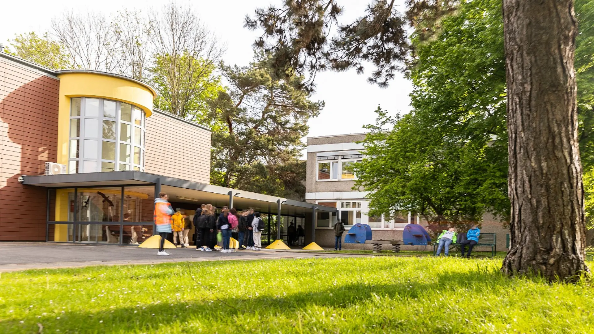  Ein gepflasterter Weg mit Leitstreifen führt durch eine Rasenfläche auf mehrere Schulgebäude zu; links befindet sich ein Backsteingebäude mit einem Spielplatz davor, in der Mitte ein helleres Gebäude mit großen Fenstern und rechts ein modernes Gebäude mit Metallverkleidung und großen Glasfronten, umgeben von Bäumen.