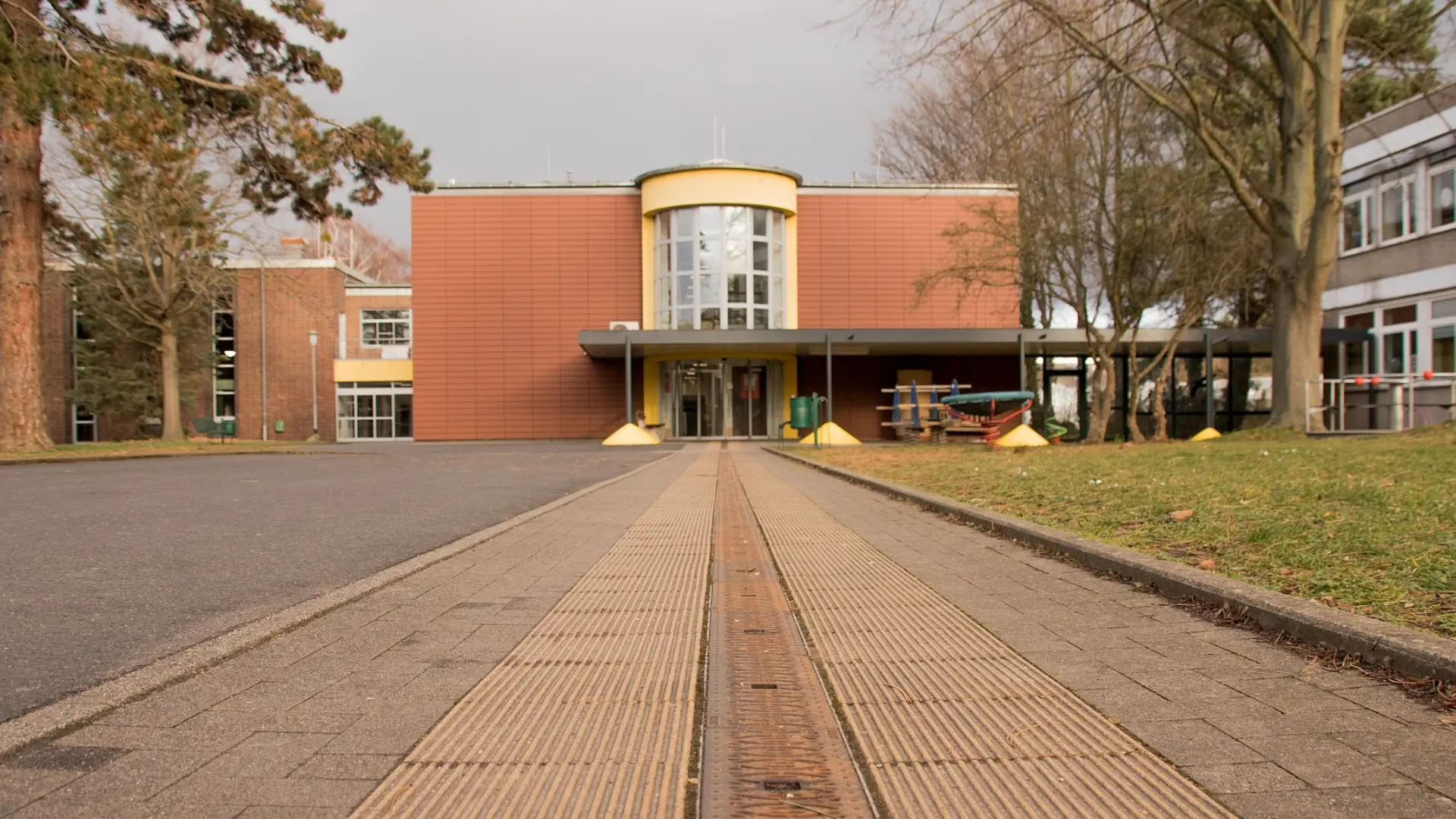  Ein breiter, gepflasterter Weg führt auf ein modernes Gebäude zu, das von Bäumen umgeben ist. In der Mitte des Weges befindet sich ein Blindenleitsystem aus gerillten Platten, das direkt zum Eingang des Gebäudes führt. Das Gebäude verfügt über eine markante, halbrunde Glasfront im oberen Bereich des Haupteingangs. Rechts und links des Weges befindet sich Rasenfläche.