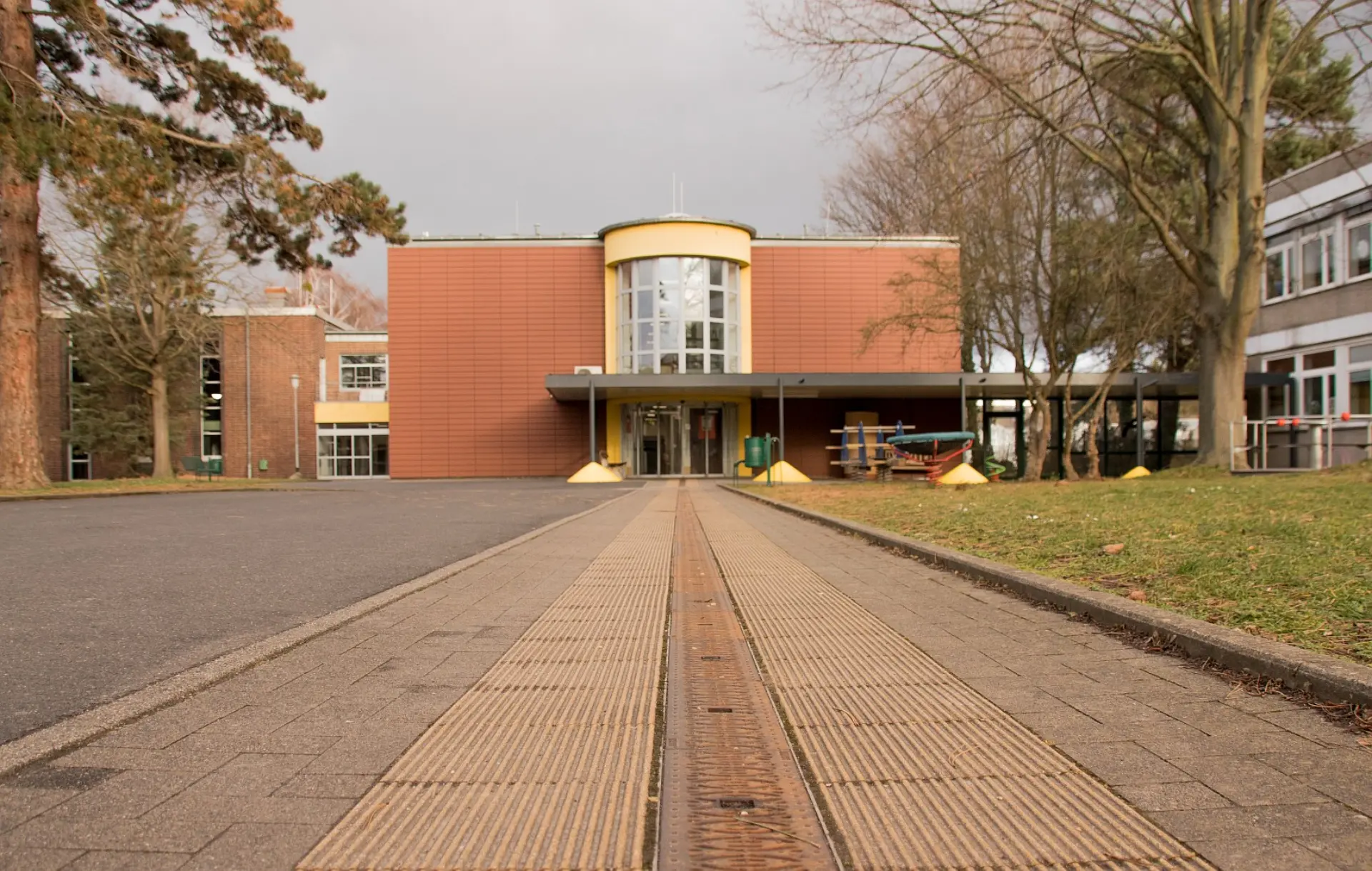 Ein breiter, gepflasterter Weg führt auf ein modernes Gebäude zu, das von Bäumen umgeben ist. In der Mitte des Weges befindet sich ein Blindenleitsystem aus gerillten Platten, das direkt zum Eingang des Gebäudes führt. Das Gebäude verfügt über eine markante, halbrunde Glasfront im oberen Bereich des Haupteingangs. Rechts und links des Weges befindet sich Rasenfläche.