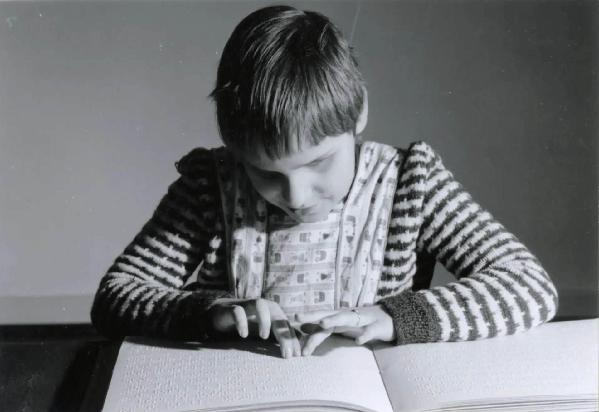 Ein monochromes Foto von einer blinden Schülerin, die am Tisch sitzt. Vor ihr ist ein Buch in Braille-Schrift, sie geht mit den Fingern darüber und liest den Text.
