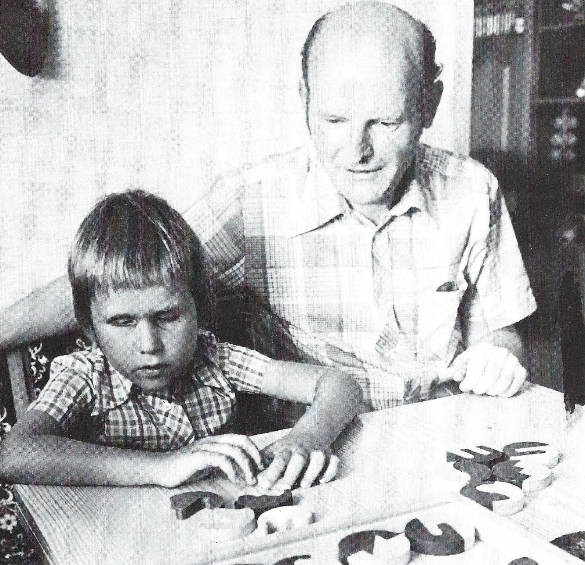 Eine alte, schwarzweiße Porträtaufnahme, die einen Mann und ein blindes Kind am Tischen sitzen zeigt. Das Kind hat vor sich Holzfiguren, die es puzzle-artig aneinander stecken kann. Zwei davon hält es in den Händen. Der Mann beobachtet von der Seite, was es tut.