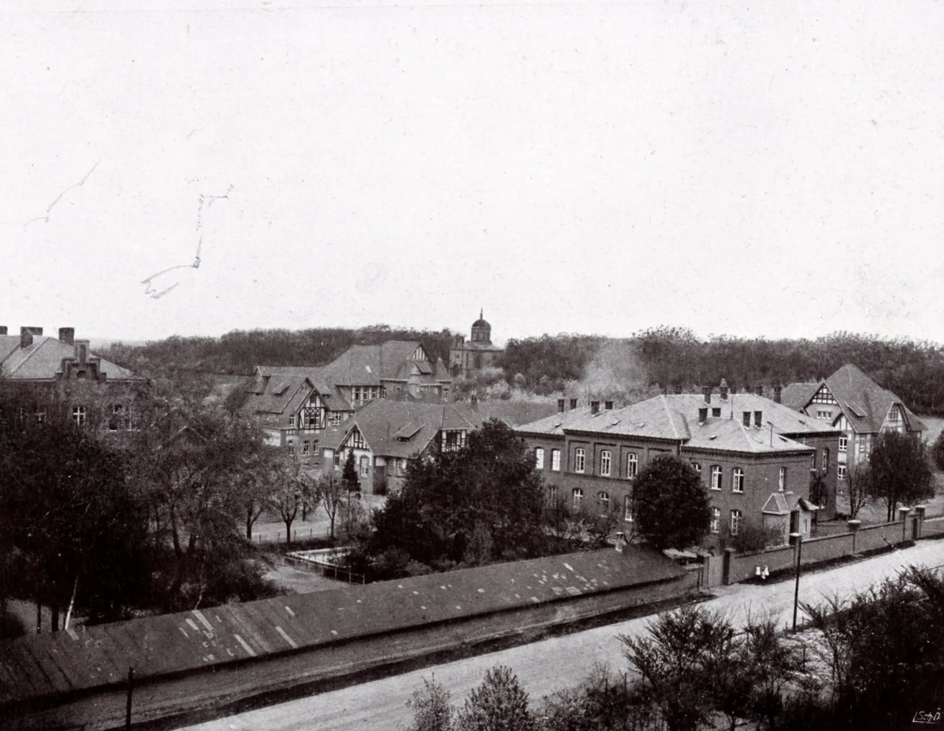 Ein historisches Schwarz-Weiß-Luftbild zeigt eine Gesamtansicht eines weitläufigen Gebäudekomplexes in einer ländlichen oder vorstädtischen Umgebung. Im Vordergrund verläuft eine Straße parallel zum unteren Bildrand, daneben befindet sich ein langes, niedriges Gebäude mit einem Schrägdach. Dahinter erstrecken sich mehrere größere Gebäude mit verschiedenen Dachformen, darunter Sattel- und Walmdächer, die von Bäumen und anderen Grünflächen umgeben sind. Im Hintergrund ist ein dichter Wald oder Baumbestand zu sehen, aus dem sich eine Kirchenkuppel oder ein Turm erhebt.