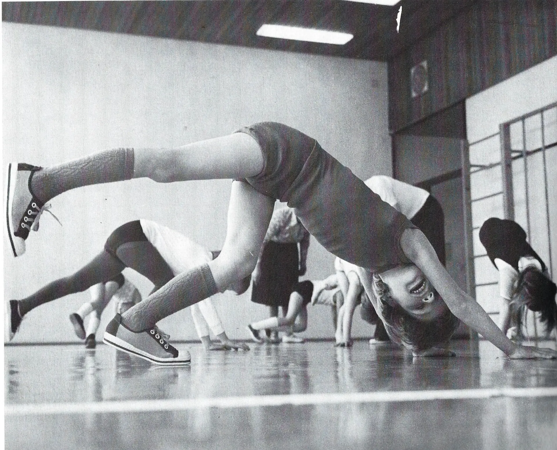  Eine Schwarzweiß-Aufnahme aus der Sporthalle der Schule. Das Foto wurde vom Boden heraus gemacht, sodass die Schüler*innen, die Gymnastik machen, aus einer tiefen Perspektive fotografiert worden und komplett zu sehen sind. Fünf Schüler*innen stehen auf aller Vieren, die Arme sind auf dem Boden nach vorne ausgestreckt, ein Bein ist auch auf dem Boden, das andere in der Luft ausgestreckt. Zwei Pädagog*innen laufen dazwischen und helfen den Kindern.