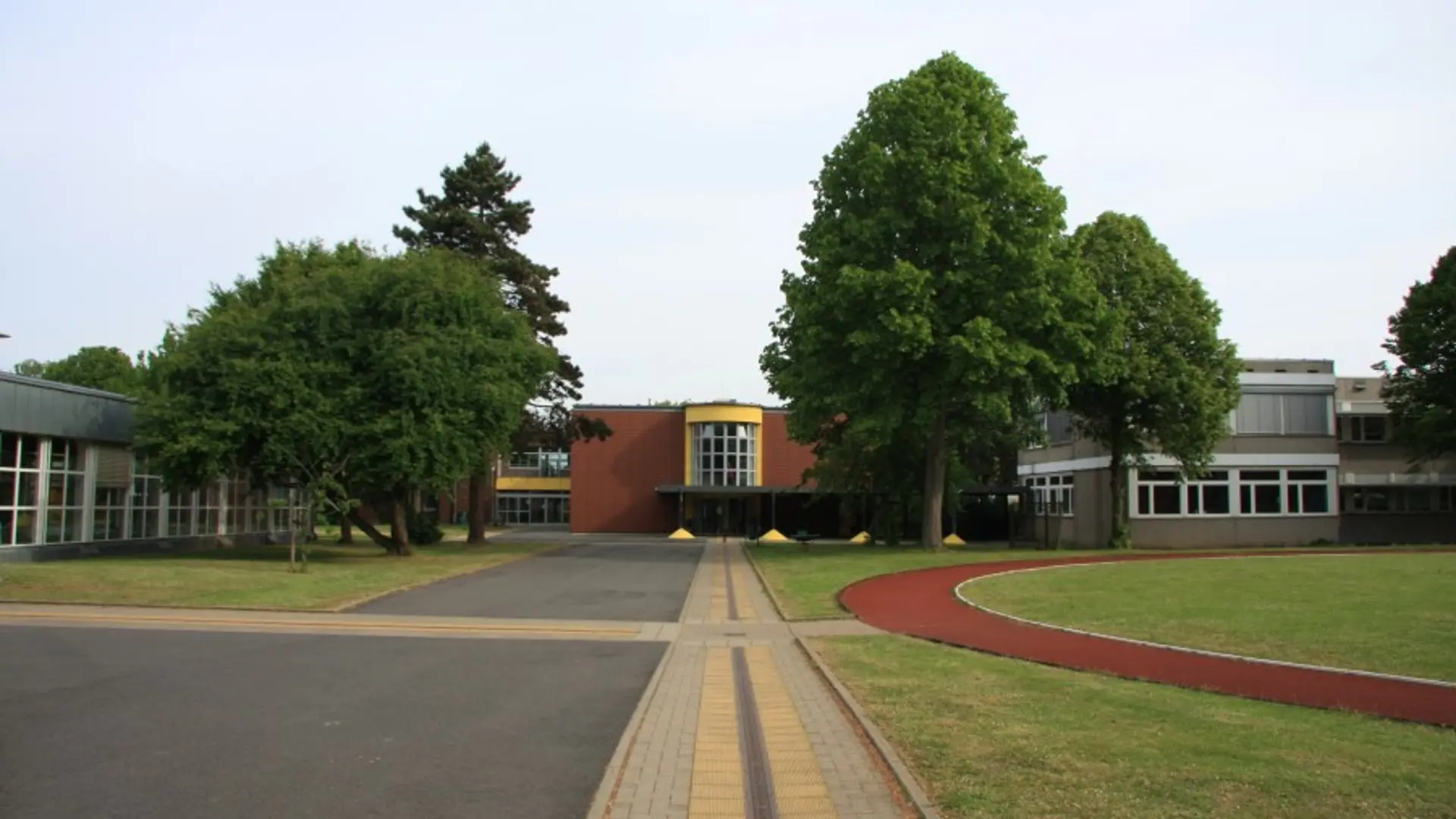 Ein Schulgebäude mit einem großen, begrünten Außenbereich an einem klaren Tag. Im Vordergrund befindet sich ein gepflasterter Weg mit Orientierungsmarkierungen für blinde Menschen, der zu einem Eingang führt. Der Weg ist von Rasenflächen gesäumt, auf der rechten Seite ist eine rote Laufbahn zu sehen. Mehrere Bäume sind auf dem Gelände verteilt. Das Gebäude selbst besteht aus mehreren verbundenen Teilen, die unterschiedliche architektonische Merkmale aufweisen, darunter große Fensterfronten und Fassaden aus Ziegeln und anderen Materialien.