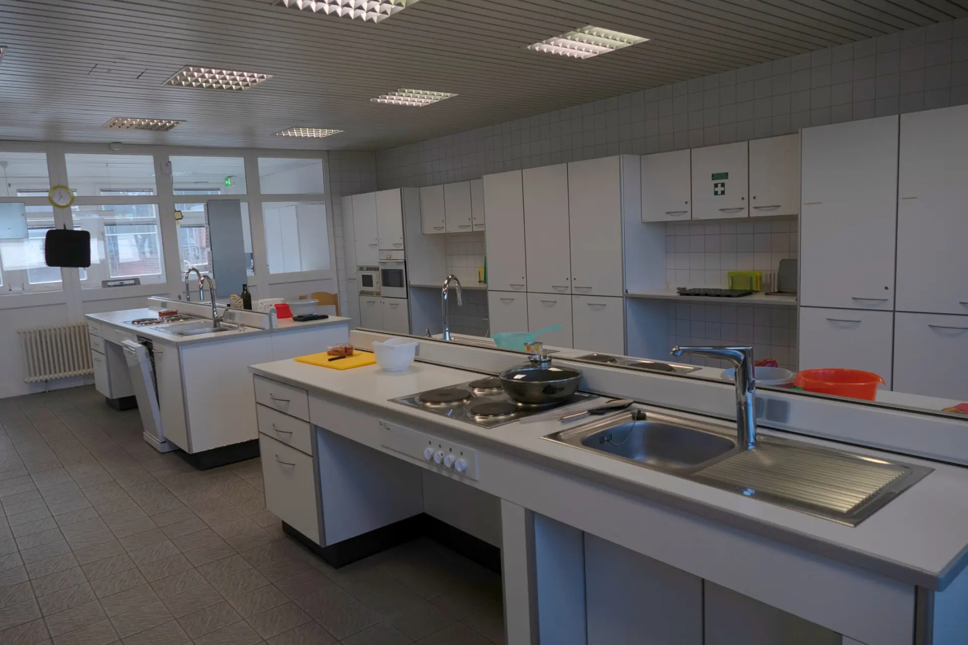 Ein gut beleuchteter, sauberer Raum, der als Lehrküche oder Hauswirtschaftszentrum dient. Es gibt mehrere weiße Küchenzeilen mit Spülbecken und Kochfeldern. An den Wänden sind viele Hängeschränke angebracht. Eine große Fensterfront auf der linken Seite lässt Tageslicht herein. Auf den Arbeitsflächen stehen verschiedene Küchenutensilien und Kochgeschirr. Der Boden ist mit Fliesen ausgelegt.