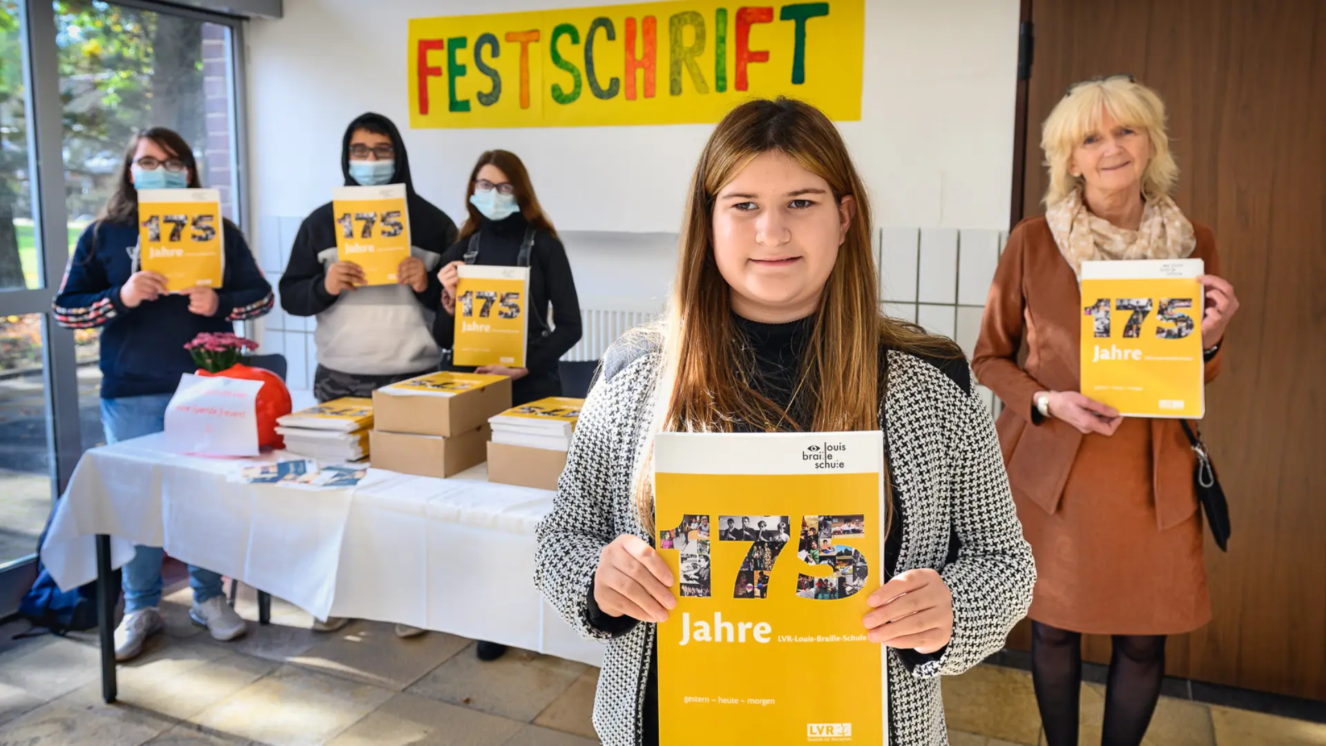  Im Flur der Schule steht ein Tisch mit Papierdecke. Vor dem Tisch steht eine Schülerin, hinter ihr (rechts im Bild) steht die Schuldirektorin Frau Grün-Klingebiel, und hinter dem Tischt im Hintergrund stehen noch drei Schüler*innen mit Corona-Masken über Mund und Nase. Alle fünf Personen halten die im DinA4-Format ausgedruckte Broschüre "175 Jahre LVR-Louis-Braille-Schule. Gestern - Heute - Morgen". Auf dem Tisch sind noch vier Kartons mit den Broschüren sowie ein Sparschwein für Spenden für die Schule beim Mitnehmen der Broschüre. An der Wand im Hintergrund klebt ein handgemachtes Plakat mit dem Wort "Festschrift".