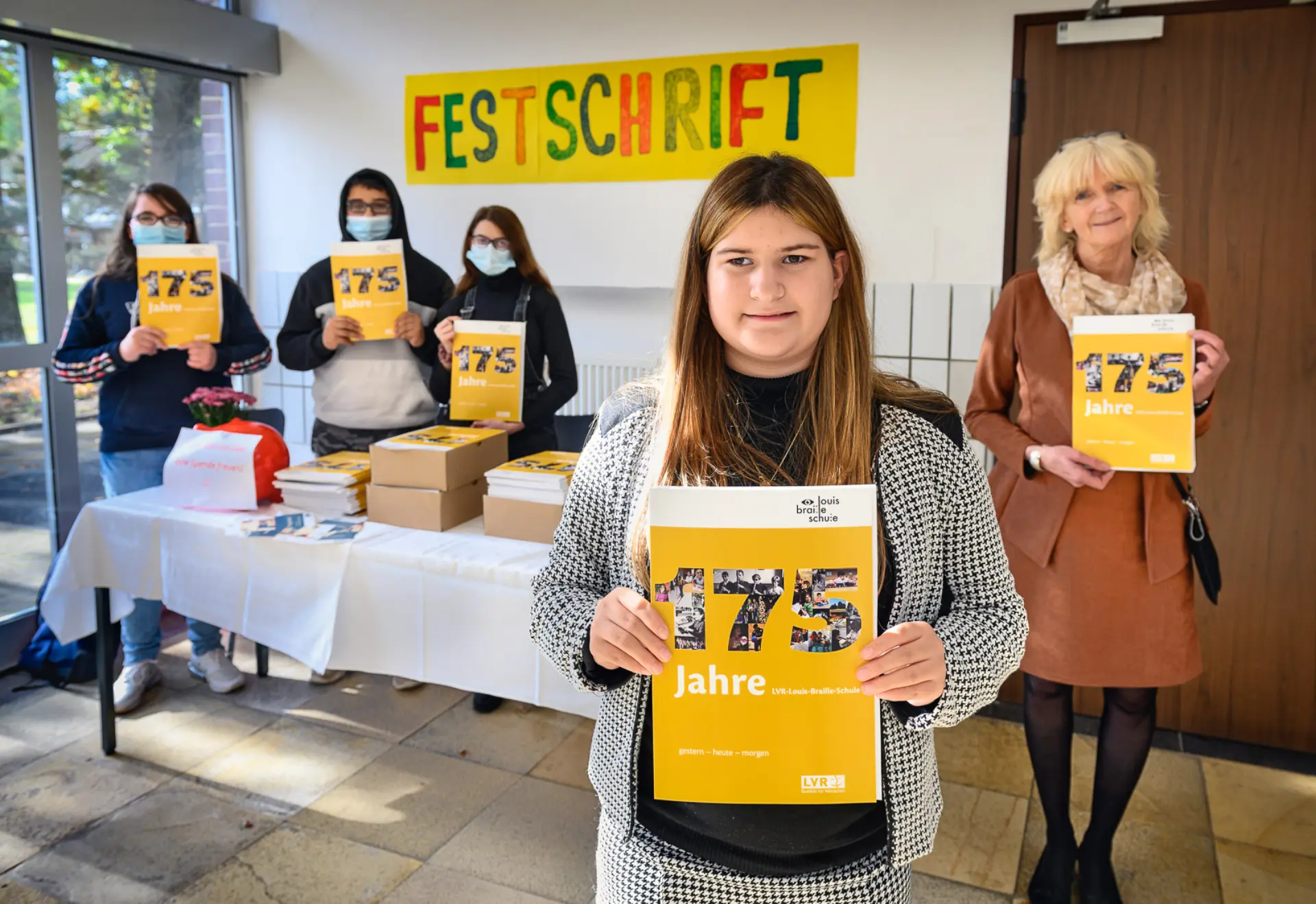 Im Flur der Schule steht ein Tisch mit Papierdecke. Vor dem Tisch steht eine Schülerin, hinter ihr (rechts im Bild) steht die Schuldirektorin Frau Grün-Klingebiel, und hinter dem Tischt im Hintergrund stehen noch drei Schüler*innen mit Corona-Masken über Mund und Nase. Alle fünf Personen halten die im DinA4-Format ausgedruckte Broschüre "175 Jahre LVR-Louis-Braille-Schule. Gestern - Heute - Morgen". Auf dem Tisch sind noch vier Kartons mit den Broschüren sowie ein Sparschwein für Spenden für die Schule beim Mitnehmen der Broschüre. An der Wand im Hintergrund klebt ein handgemachtes Plakat mit dem Wort "Festschrift".