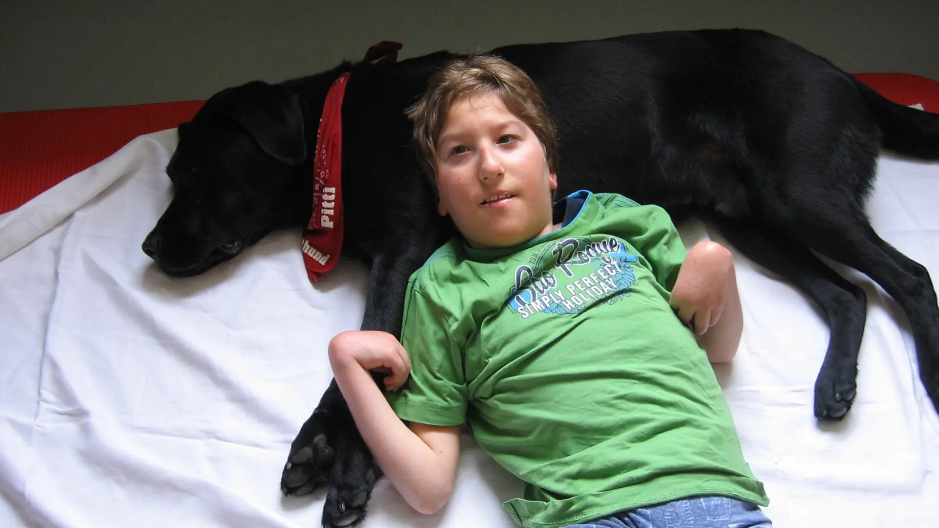  Ein Schüler mit komplexer Beeinträchtigung liegt auf einer Decke und einem Bettlacken auf dem Boden. Sein Kopf liegt bequem auf der Brust eines seitlich liegenden Hundes, der entspannt die Augen zuhat. Das ist ein Therapiehund und er hat ein Halstuch am Hals gebunden. Darauf steht der Name "Pitti".
