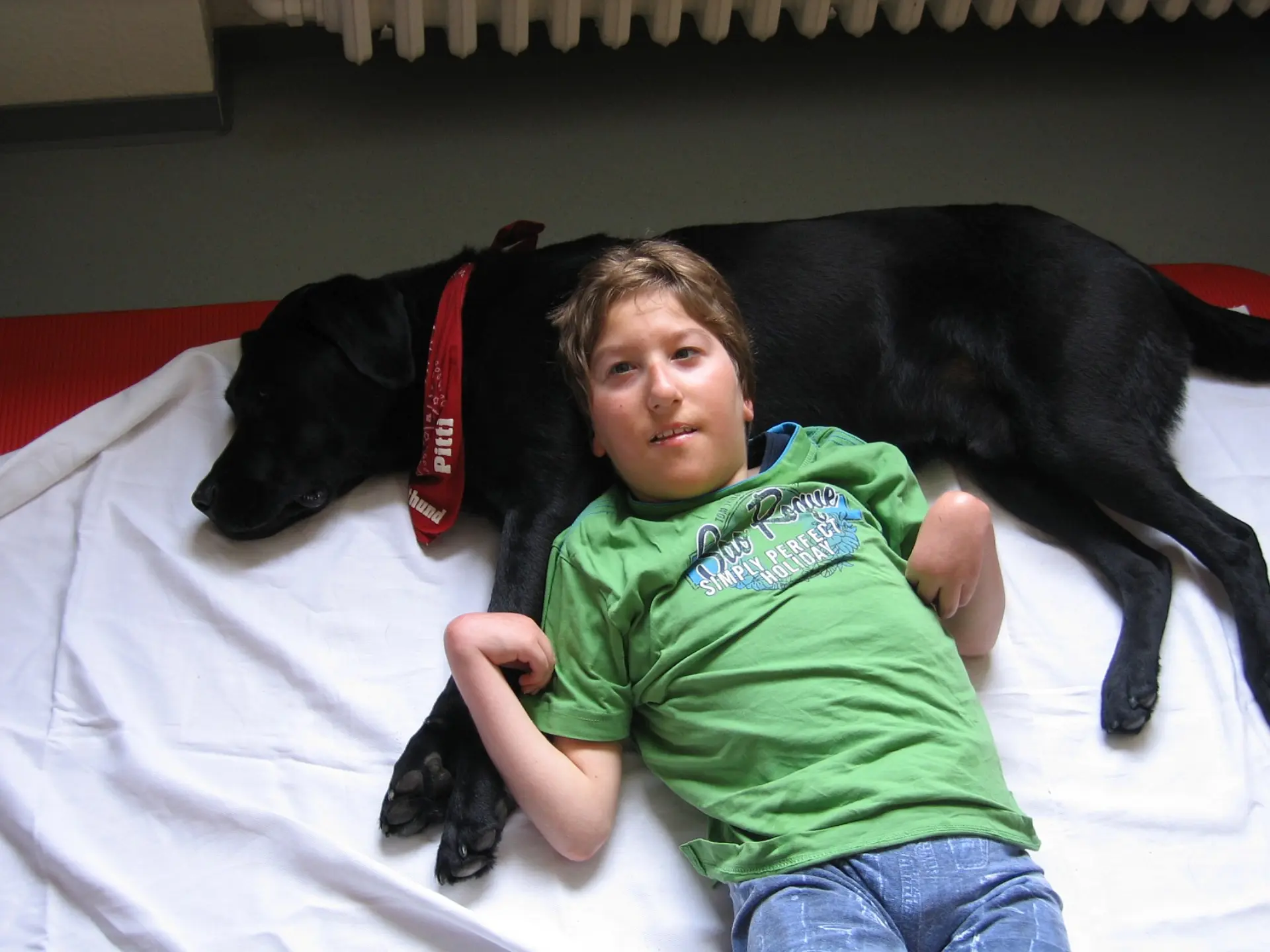 Ein Schüler mit komplexer Beeinträchtigung liegt auf einer Decke und einem Bettlacken auf dem Boden. Sein Kopf liegt bequem auf der Brust eines seitlich liegenden Hundes, der entspannt die Augen zuhat. Das ist ein Therapiehund und er hat ein Halstuch am Hals gebunden. Darauf steht der Name "Pitti".