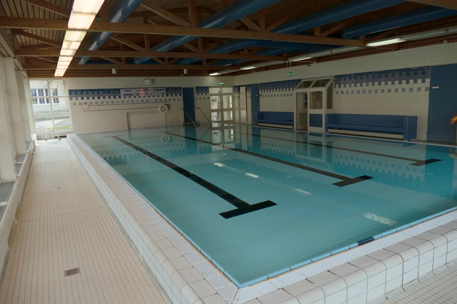 Ein Schwimmbad-Innenraum, von einer erhöhten Position aus fotografiert. Das Becken ist mit klarem Wasser gefüllt und hat zwei dicke Linien, die vom Startblock bis zum anderen Ende verlaufen. Die Wände sind gefliest. Im Hintergrund sind Fenster, Bänke und Türen zu sehen. Die Decke besteht aus Holzbalken.