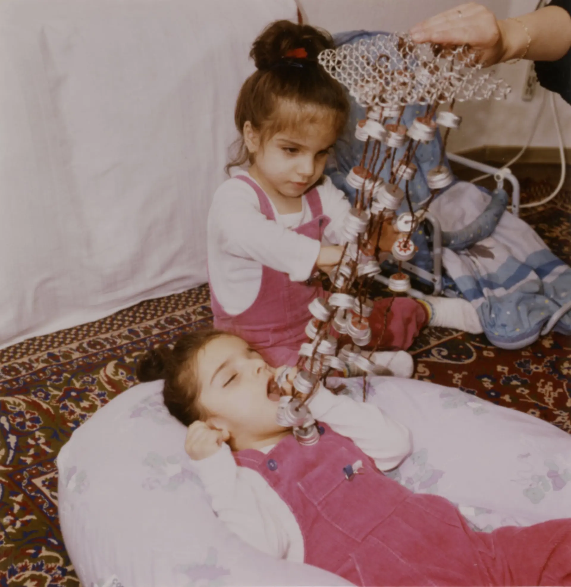 Zwei Mädchen mit komplexer Beeinträchtigung befinden sich auf dem Boden, der mit einem detailreichen Teppich bedeckt ist. Vom Aussehen her könnte man sagen, dass sie Zwillinge sind. Das eine Mädchen liegt auf einem großen Kissen, die Arme sind zu ihrem Kopf gestreckt, es hat den Mund offen. Die Hand einer erwachsenen Person hält eine Art Spielmobile mit vielen kleinen Kügelchen über dem Kind. Das zwei Mädchen sitzt auf dem Boden neben dem anderen und greift zu den Kügelchen.